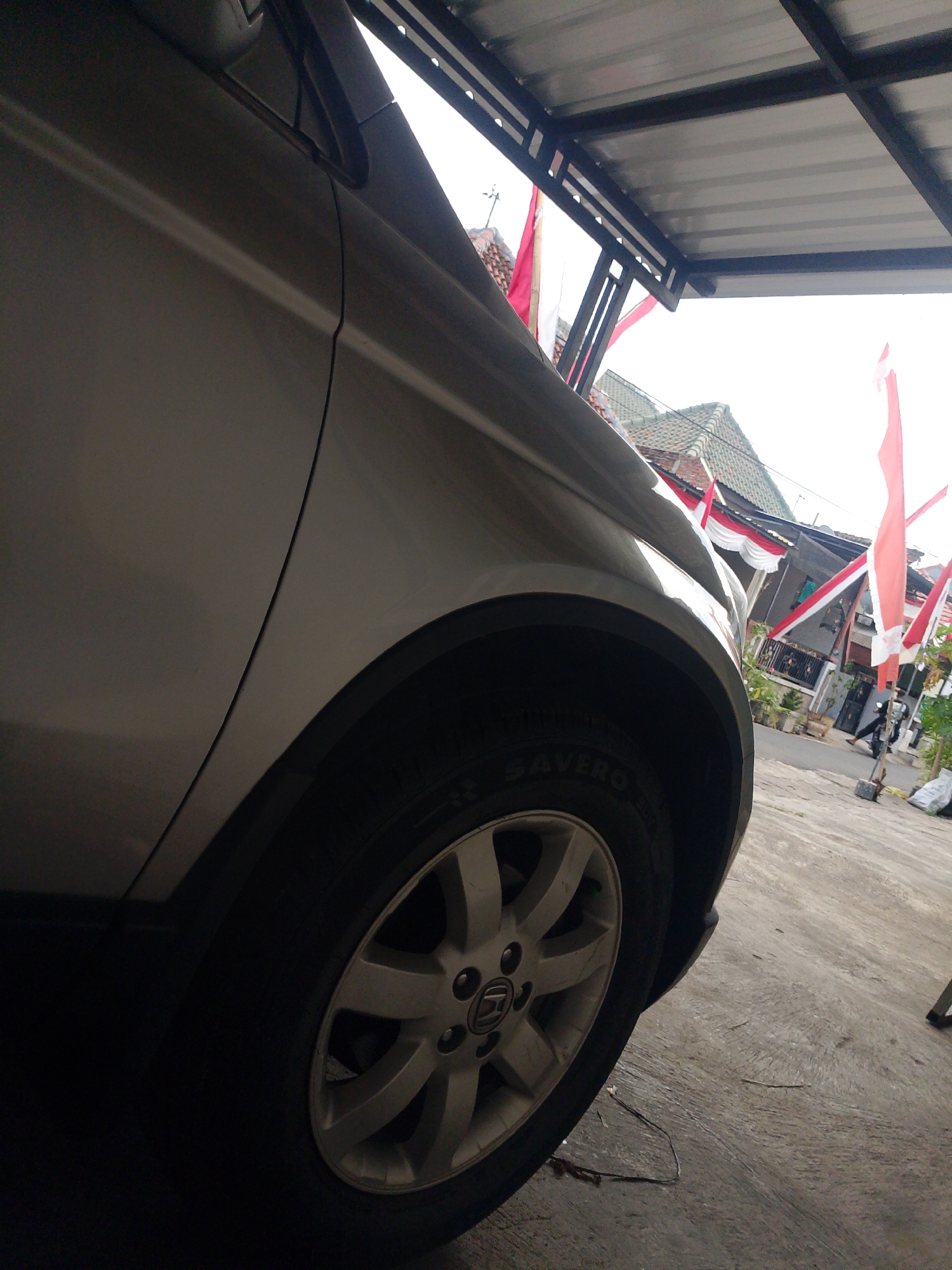 The image is an exterior, eye-level, medium shot of the front fender and wheel of a silver car parked under a metal awning. The car is angled towards the right, with its side taking up the majority of the left side of the frame. The wheel, with a silver alloy rim, is visible beneath the fender. The tire has text that reads "SAVERO". The awning above casts a shadow over the car and the ground.
Behind the car, a street scene is visible through the gap in the awning. Red and white flags, commonly used for Indonesian Independence Day celebrations, are hung along the street and on houses. The houses appear to be typical Indonesian residential structures with tiled roofs. There are potted plants in front of some houses.
Further down the street, a person is walking away from the camera towards the right. The ground is paved with concrete. The overall lighting suggests it is daytime, possibly overcast. The context of the Indonesian flags strongly suggests the image was taken in Banyuwangi, Indonesia, during a period of patriotic celebration.