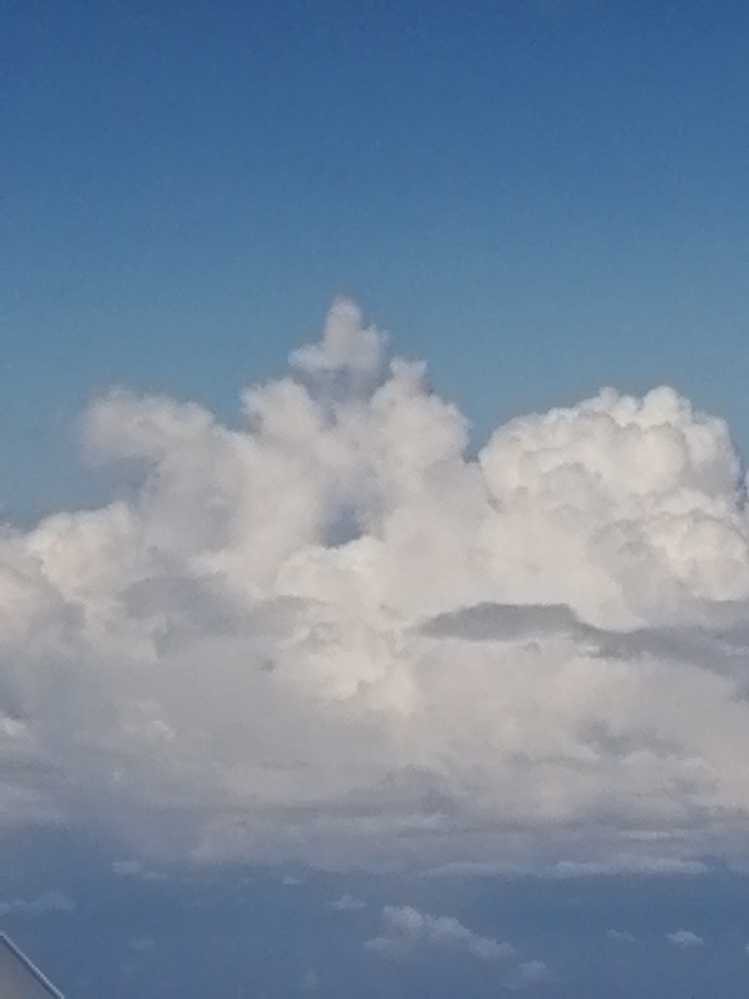 The image depicts a cloudy sky with a bright blue background. The clouds are predominantly white and appear to be cumuliform in shape, exhibiting varying degrees of density and texture. A specific cloud formation in the center has a distinct, irregular peak. The lower portion of the image reveals a hazy gradient, suggesting lower-lying atmospheric layers or distant features.