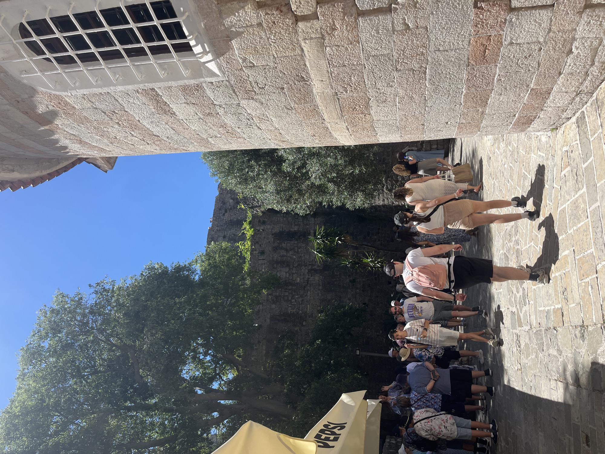 The image shows a narrow street in the city of Budva, Montenegro. The street is paved with stone blocks. On either side are buildings constructed with beige stone blocks, one of them with a gated window on the top. The walls cast shadows on the stone path.
A line of people are walking away, while another group of people is queuing to the right of the frame next to a beige Pepsi marquee.
Beyond the buildings, there is a stone wall, trees and blue sky.