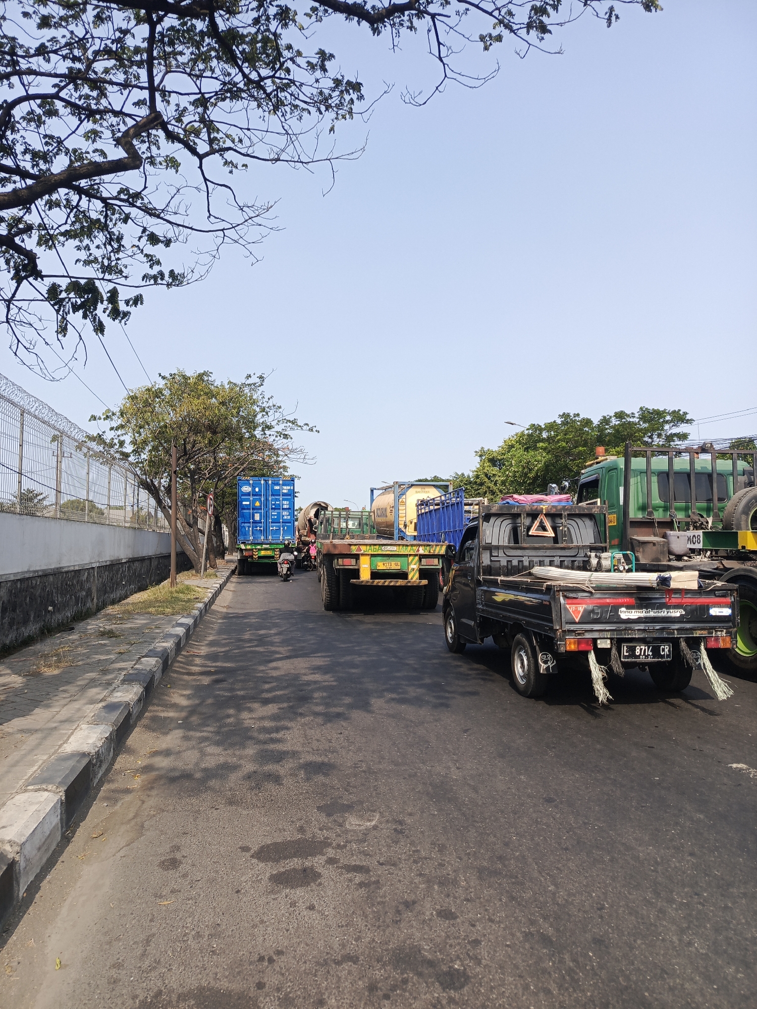 The image shows a street scene in Surabaya, Indonesia, with several trucks and a utility vehicle on a paved road. The sky is clear and blue, with some tree branches visible in the upper portion of the frame. On the left side of the road, there is a sidewalk with a concrete curb and a wall topped with a chain-link fence. Beyond the wall, trees and vegetation are visible.
The vehicles in the scene include:
* A blue shipping container on a truck, with a motorcycle passing behind it.
* A yellow tanker truck.
* A black utility vehicle with metal pipes and other materials in its bed. It has a license plate that reads "L 8714 CR".
* A green truck with a large cylindrical object on its back.
The overall impression is one of an industrial or commercial area, likely during the daytime, with traffic on the road.