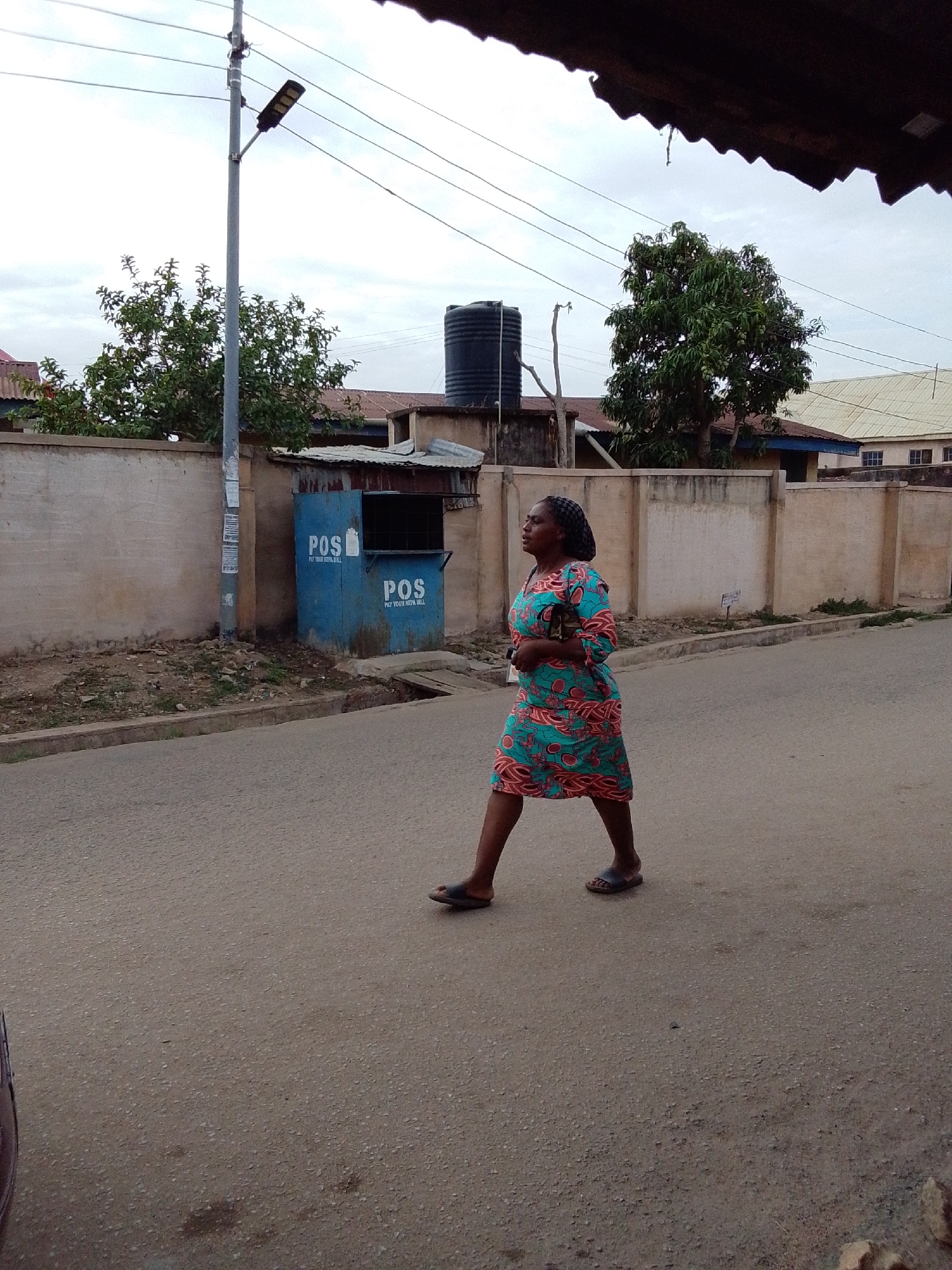 The image presents an outdoor street scene in Jos, Nigeria, under an overcast sky. The main subject is a woman walking on a paved road. She has braided hair and is wearing a colorful midi-length dress with a teal/blue base and intricate orange and pink patterns. She is wearing flip-flops and appears to be carrying a small item in her hand.
In the background, to the left, a tall grey utility pole with a black, solar-powered street light stands prominently, with several power lines stretched across the frame. Below the pole, behind a low concrete wall, is a lush green tree. Adjacent to the pole and the wall is a small, blue, box-like kiosk. The kiosk has "POS" written in white lettering on its side twice, along with the text "PAY YOUR NEPA BILL," indicating it serves as a point-of-sale service, likely for utility payments.
Further into the background, parts of houses with corrugated roofs and a large black water tank are visible. Another green tree is seen towards the right side of the background. The road surface is asphalt, and a narrow dirt strip lies between the road and the concrete wall. The setting suggests a modest residential or commercial area.