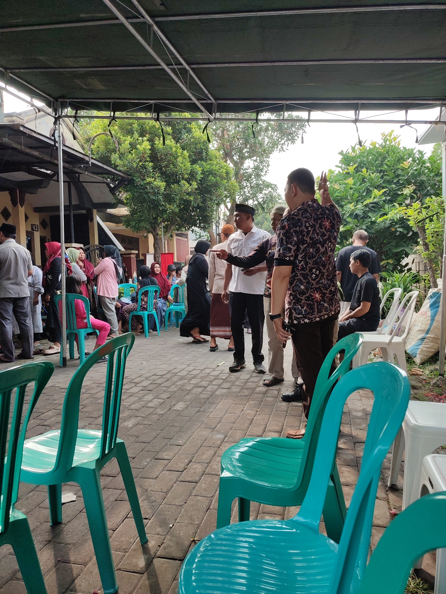 A group of people are gathered under an outdoor canopy in what appears to be a residential area in Muktisari, Indonesia. The canopy is supported by metal poles and covered with a dark green tarp. The ground is paved with interlocking bricks. In the foreground, several bright turquoise plastic chairs are arranged, some facing away from the camera. In the midground, a diverse group of individuals, men and women dressed in traditional Indonesian clothing such as sarongs and hijabs, are standing and sitting. Some are engaged in conversation, while others appear to be observing or waiting. One man in a patterned shirt and dark pants is gesturing with his hand. Behind the group, a building with beige walls and dark trim is visible, along with lush green trees and foliage. The overall scene suggests a community gathering or event.