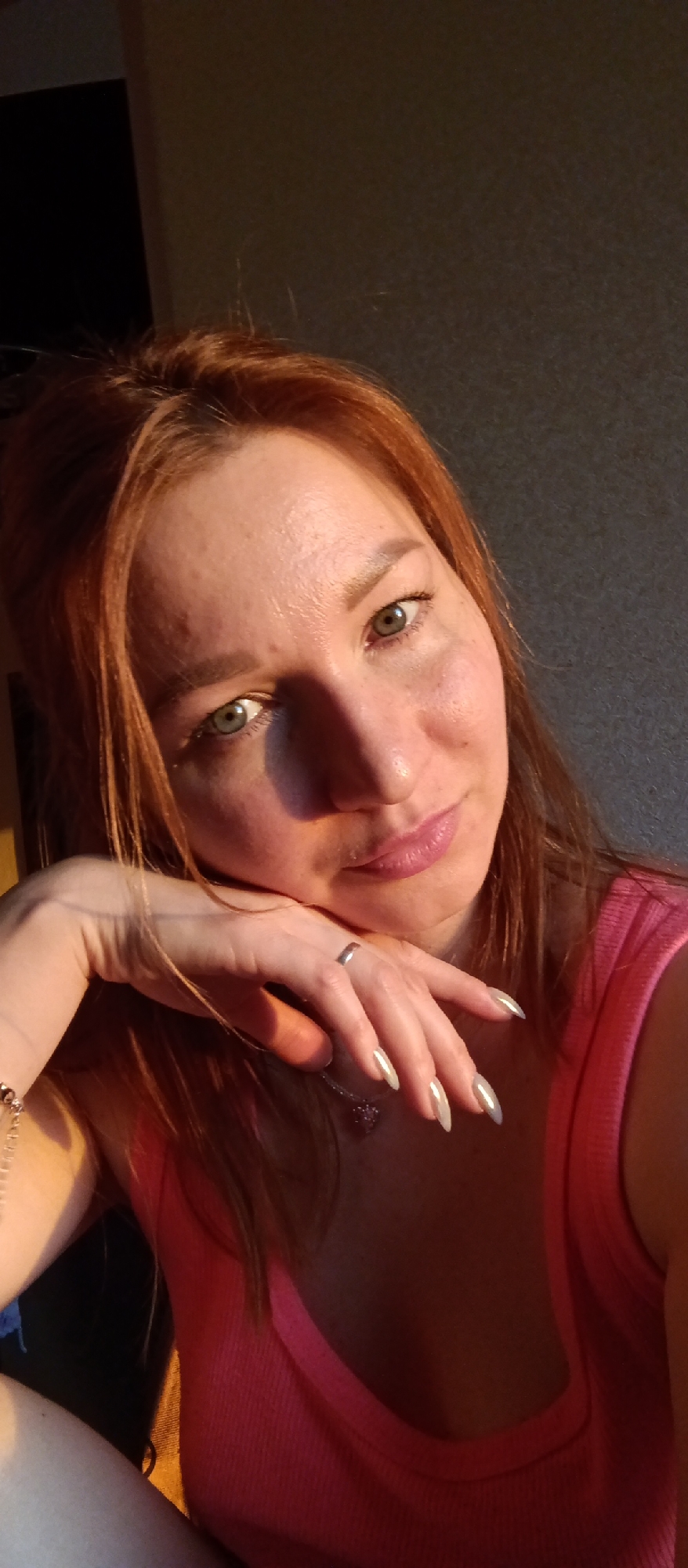 A close-up indoor photograph shows a Caucasian woman with fair skin, shoulder-length auburn hair, and green eyes. She is wearing a pink tank top. Her left hand is positioned with her index finger resting on her chin, and she is wearing a silver ring on that finger. She is also wearing a silver chain necklace with a heart pendant. Her fingernails are long and almond-shaped, with a light-colored polish. The background is a plain light gray wall, and there is a dark object, possibly a door, visible in the upper left corner. The lighting appears to be natural, coming from the right side of the frame, casting shadows across her face and hand. The location is Unknown, Unknown.