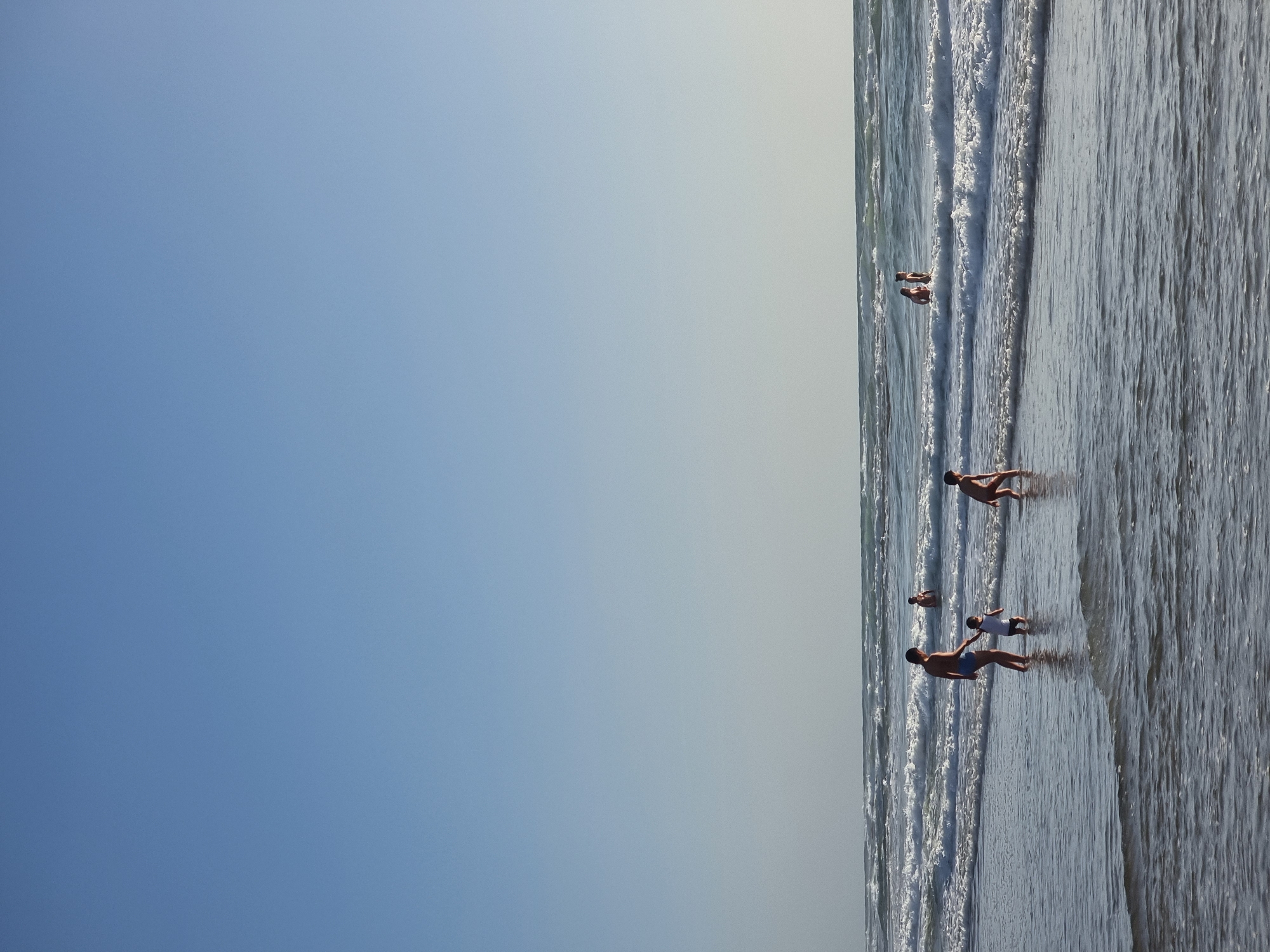 The image shows a beach scene, likely in Tel Aviv, Israel. On the right side of the frame is the sea, with waves washing onto the sandy shore. Several people are in the water, some are swimming while others are standing closer to the beach. The left side of the image is a clear blue sky. The horizon line is distinct.