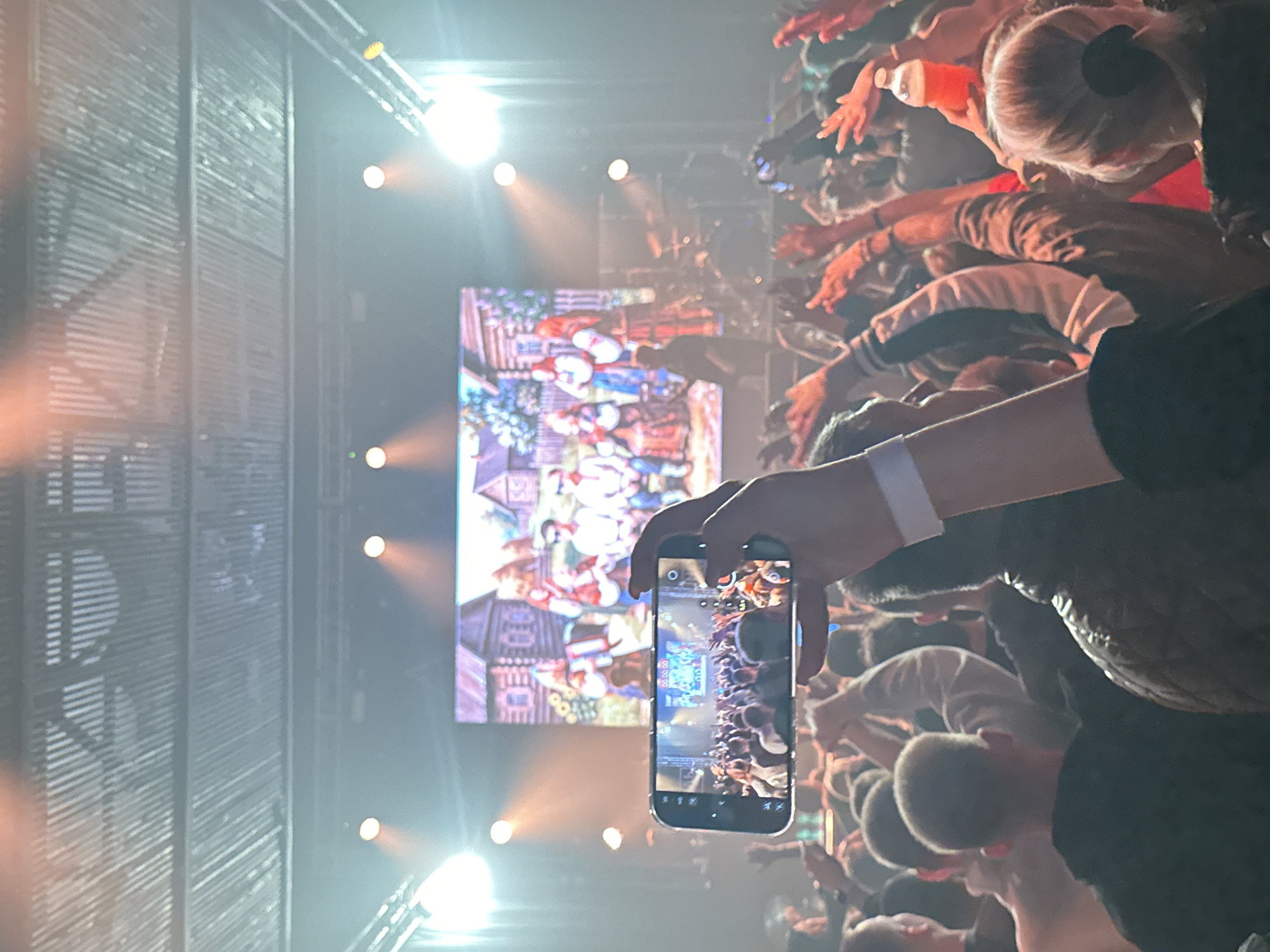 A photograph depicts a crowd of people at a concert in Izhevsk, Russia. The crowd is dense, with many people raising their arms. A large screen displays an image of artwork. A person in the foreground holds up a smartphone, capturing a photo or video of the concert. Stage lighting is visible. The venue is indoors.