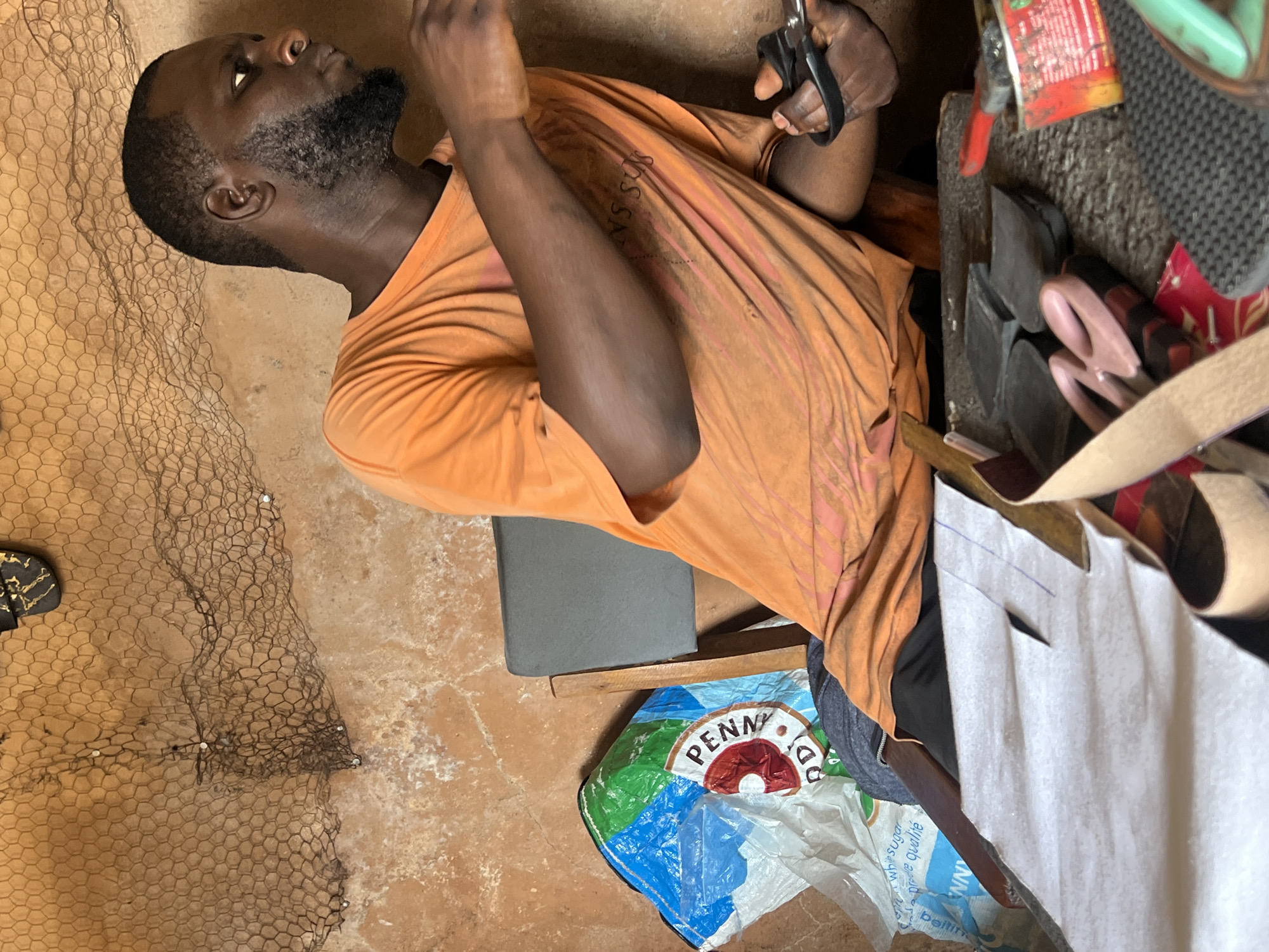 In a dimly lit, rustic workshop, a Black man with a short beard and wearing an orange t-shirt is actively engaged in crafting a shoe. He is seated, leaning forward, his right hand holding a pair of black-handled scissors, which he uses to cut a piece of white paper. In front of him lies a large sheet of white paper with blue markings, indicating a pattern or design.
The workshop's walls are made of a rough, textured material, possibly mud or concrete, with a dark, hexagonal-mesh net hanging from the ceiling. A large, multi-colored plastic bag with faded lettering, possibly a food sack, is placed on the floor to the man's left. The floor appears to be a rough, uneven surface.
On a workbench in front of the man, various shoe-making tools and materials are scattered, including several small, dark, rectangular pieces of material, possibly sole components. A pink-handled pair of scissors, a wooden ruler, and a small red and yellow container are also visible. The overall impression is of a functional, albeit somewhat cluttered, workspace dedicated to footwear production. The scene suggests a local craft setting, possibly in Jos, Nigeria, given the context.