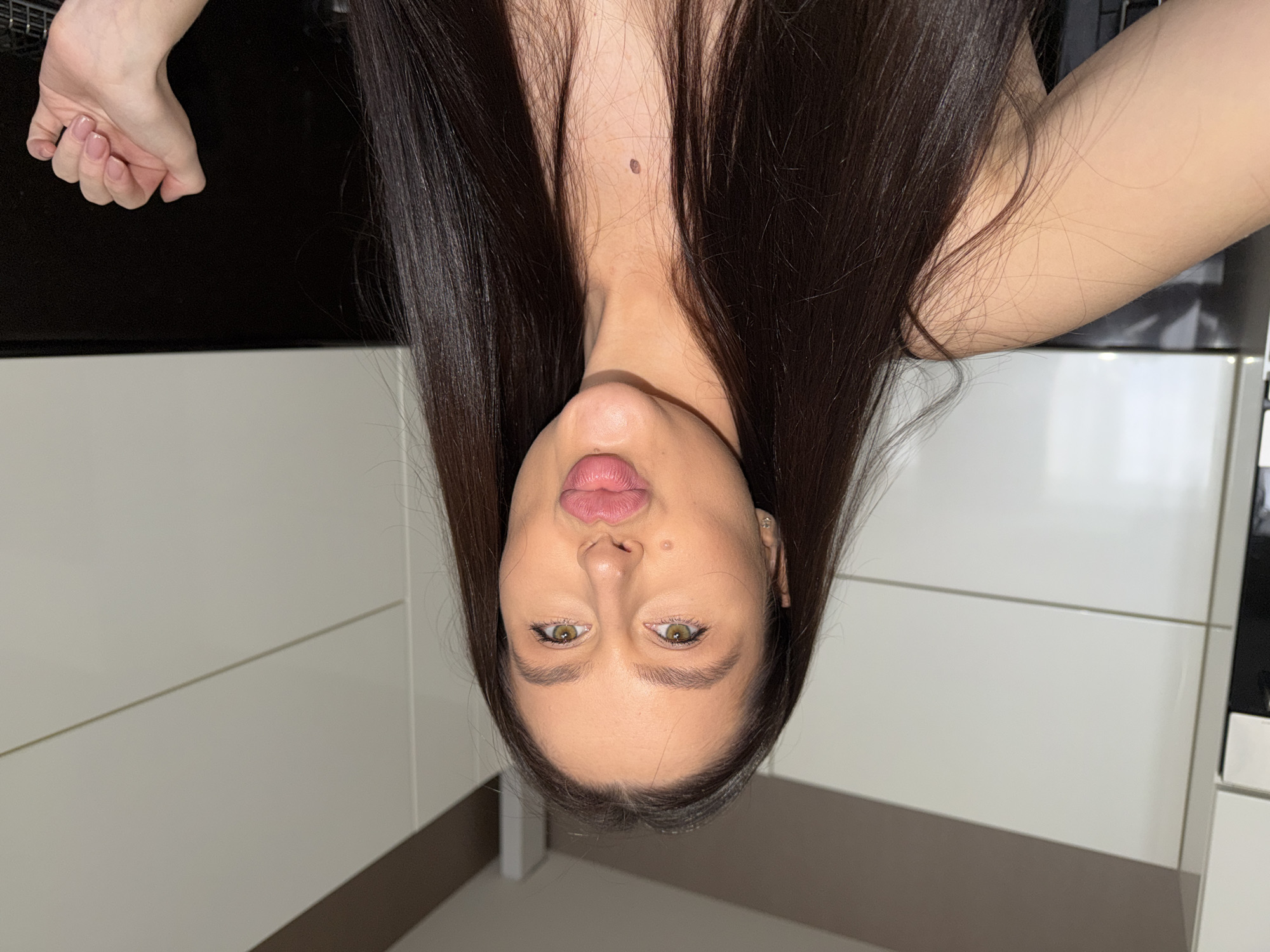 An indoor portrait features a young woman with long, dark, straight hair, captured in a close-up shot from an inverted perspective, which suggests she is looking down into the camera. She has fair skin, prominent greenish-brown eyes, and full, pink lips that are slightly pursed. A small mole or beauty mark is visible below her right eye, and another mole is present on her upper chest/neck area, just below the frame of her chin. Her left hand, with neatly manicured, light-colored nails, is visible in the upper left corner of the frame. Part of her bare shoulder and arm are also visible on the right side of the image.
The background consists of a dark, reflective surface immediately behind her head, possibly a countertop or a dark piece of furniture. Below this, the setting transitions to modern, light-colored, reflective panels or cabinets, suggesting a contemporary kitchen or living space. A thin horizontal line is visible across these panels, indicating a joint or design element. A glimpse of what appears to be a black keyboard or similar electronic device can be seen in the far upper right corner near her shoulder. The lighting is artificial, illuminating her face and the immediate surroundings.
The woman is posing for the camera, her direct gaze and slightly pursed lips conveying a neutral to mildly engaging expression. There is no discernible text in the image. The scene takes place indoors, likely in an apartment or house located in Khimki, Russia.