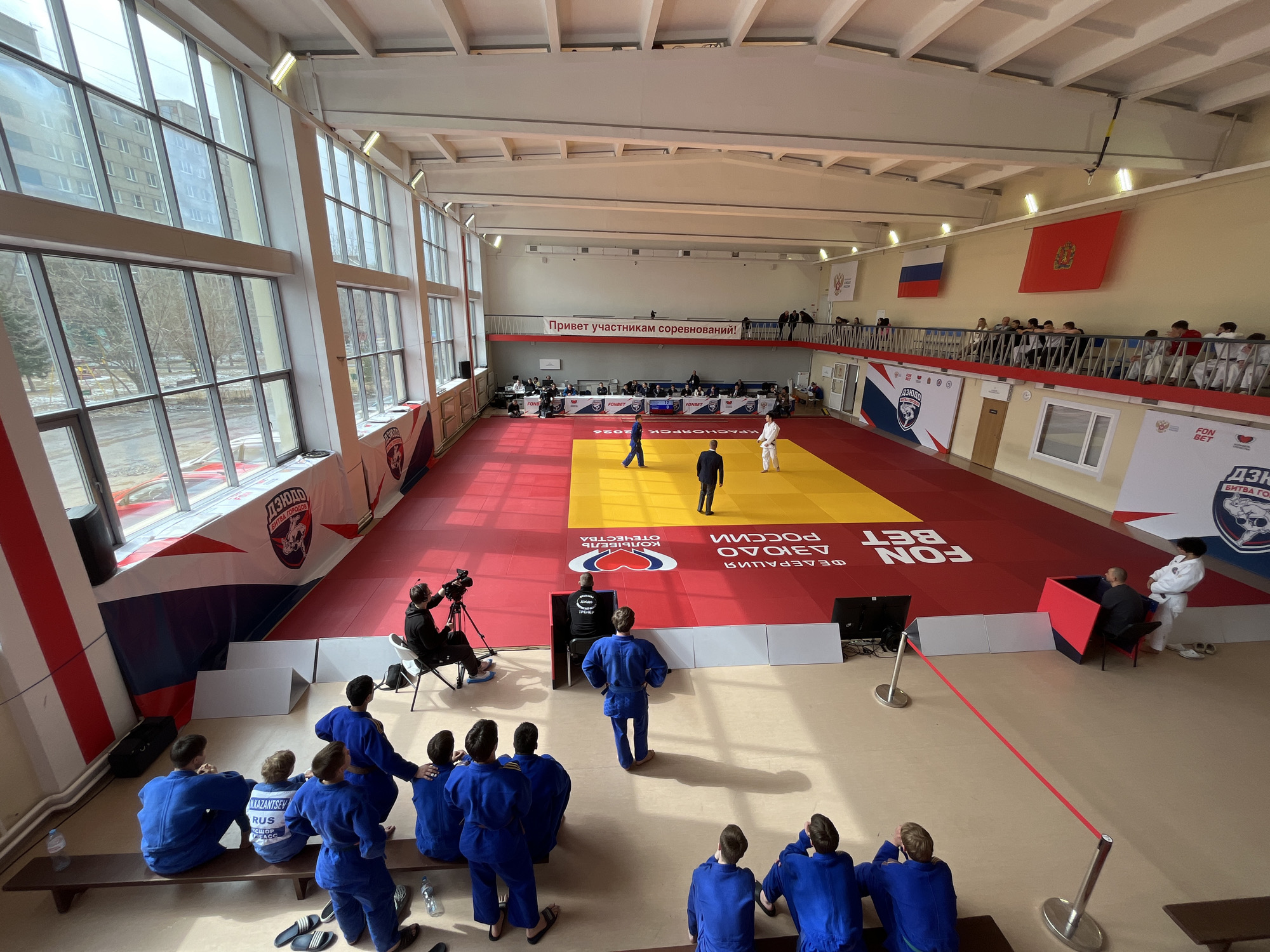 The image shows a judo competition in Krasnoyarsk, Russia. The judo mat is red and yellow. Two judokas are on the mat. A referee is in the center of the mat. A cameraman is filming the competition. A group of judokas in blue judogis are watching the competition. A red rope separates the competitors from the audience. A banner reads, "Welcome participants of the competition!" Russian and Krasnoyarsk flags are displayed on the wall. Sponsor banners for FONBET and дзн are placed around the mat. A sign indicates "MKAZANTSEV RUS СШОР FACC". The environment is a gymnasium or sports hall. Large windows are present, and a balcony overlooks the mat.