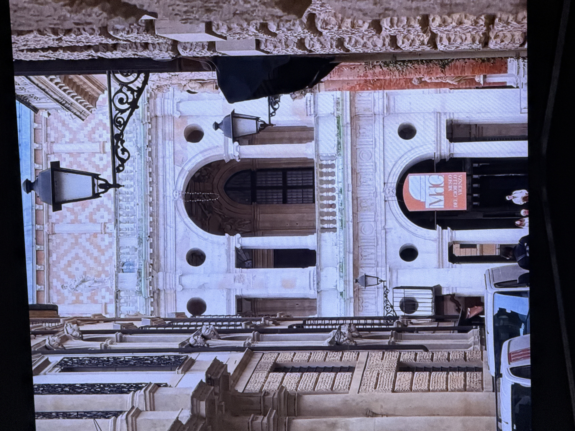 The image depicts a street scene with ornate architecture, featuring classical elements like columns, arches, and decorative stonework. Two white vans are parked in the foreground, partially obscuring a street entrance. A banner hanging above the entrance reads "MGC MUSEO DEL GIOIELLO VICENZA", indicating it is a museum. A few people are visible near the entrance and on the street. The buildings are elaborately decorated with statues and intricate patterns on their facades. Street lamps are mounted on ornate brackets on the buildings. The lighting suggests it is daytime, with some areas in shadow. The location is identified as Vicenza, Italy, not Dubai.