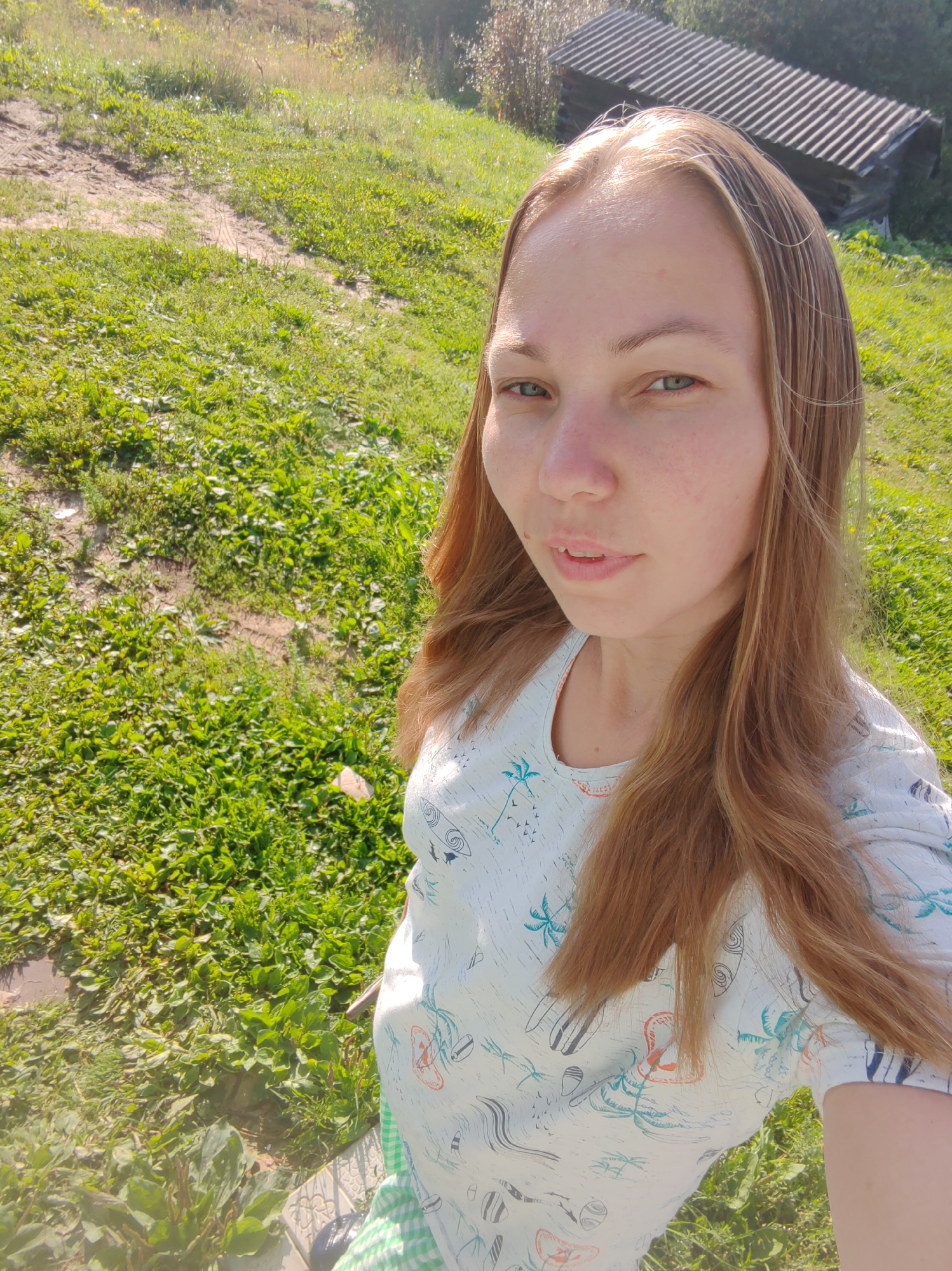 A young woman with shoulder-length blonde hair and green eyes is captured in a selfie. She is outdoors, in a grassy, uneven terrain with some dry patches and tire tracks. In the background, a wooden structure with a corrugated roof is visible on a slope. The woman is wearing a white t-shirt with a tropical print featuring palm trees and surfboards, and a green and white checkered garment, possibly shorts or a skirt, is visible at the bottom of the frame. The lighting suggests it is a sunny day. The location is identified as Чухлэм, Russia.