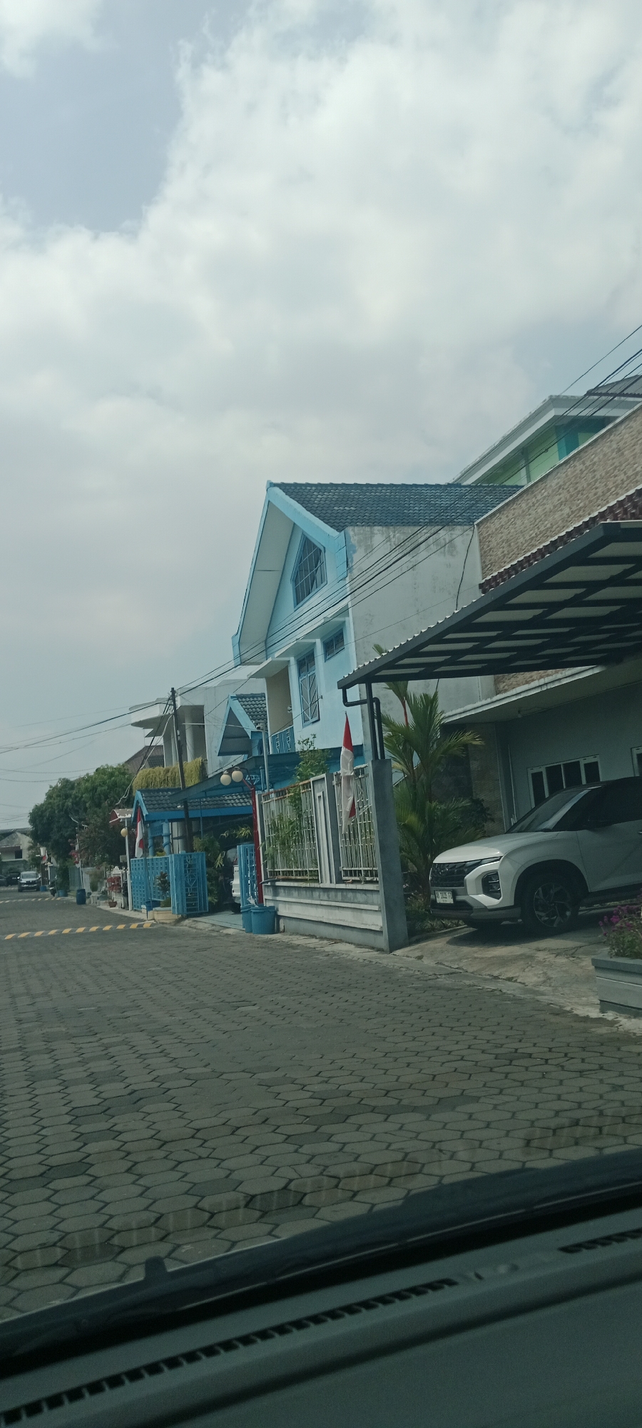The image depicts a street scene in Banyuraden, Indonesia, on an overcast day. The foreground shows a paved road with hexagonal interlocking concrete blocks. A portion of a car's dashboard is visible at the very bottom of the frame. To the right, a light-colored SUV is parked under a covered carport. The background features a row of houses and buildings. One prominent house is light blue with a pitched dark roof and distinctive triangular window accents. Another building to the right has a textured beige facade and a dark-covered awning. Several Indonesian flags are visible, attached to poles and decorative elements in front of the houses. Green trees and foliage are present along the street. The sky is filled with scattered clouds. No people are clearly visible in the scene.
