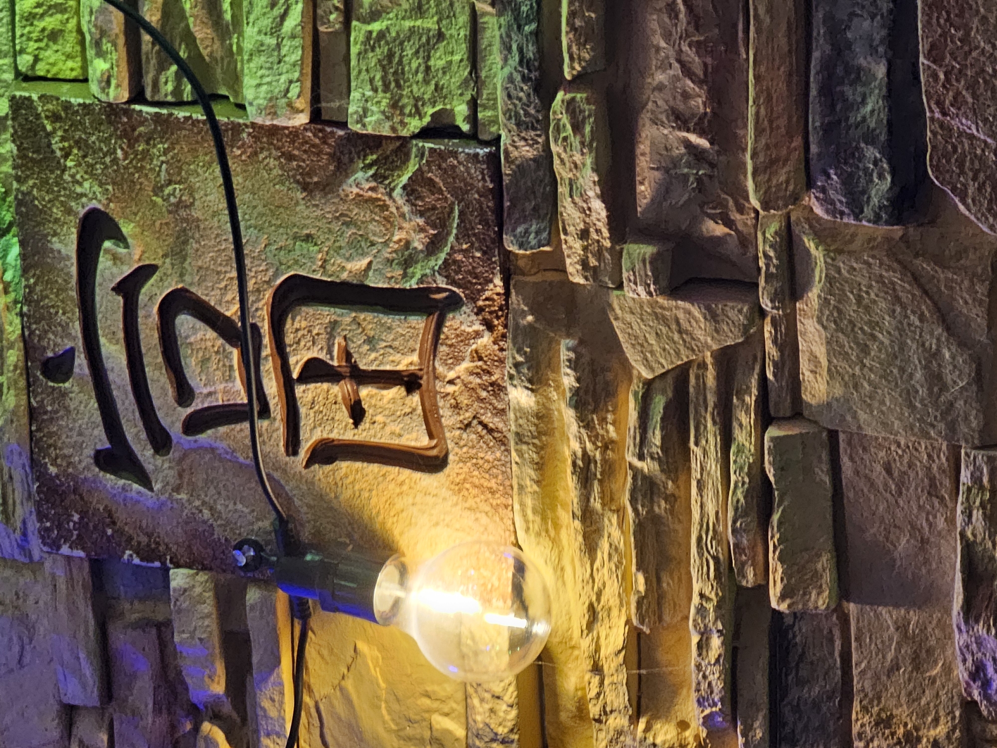 This media file shows a close-up view of a stone wall adorned with dark, stylized characters. The wall itself is constructed from irregularly shaped, textured stones, illuminated by a combination of warm, golden light and cooler green and purple hues, suggesting artificial lighting. A black wire curves across the stone surface, leading to a clear, Edison-style light bulb that is illuminated, casting a bright glow on the wall and the characters. The characters appear to be in an East Asian script, possibly Chinese or Japanese. The overall impression is one of an atmospheric, perhaps dimly lit, indoor space with decorative elements. No people are visible. The time of day is indeterminate due to the artificial lighting. No weather or specific location cues are apparent within the frame itself, other than the provided context of Skadovsk, Ukraine.