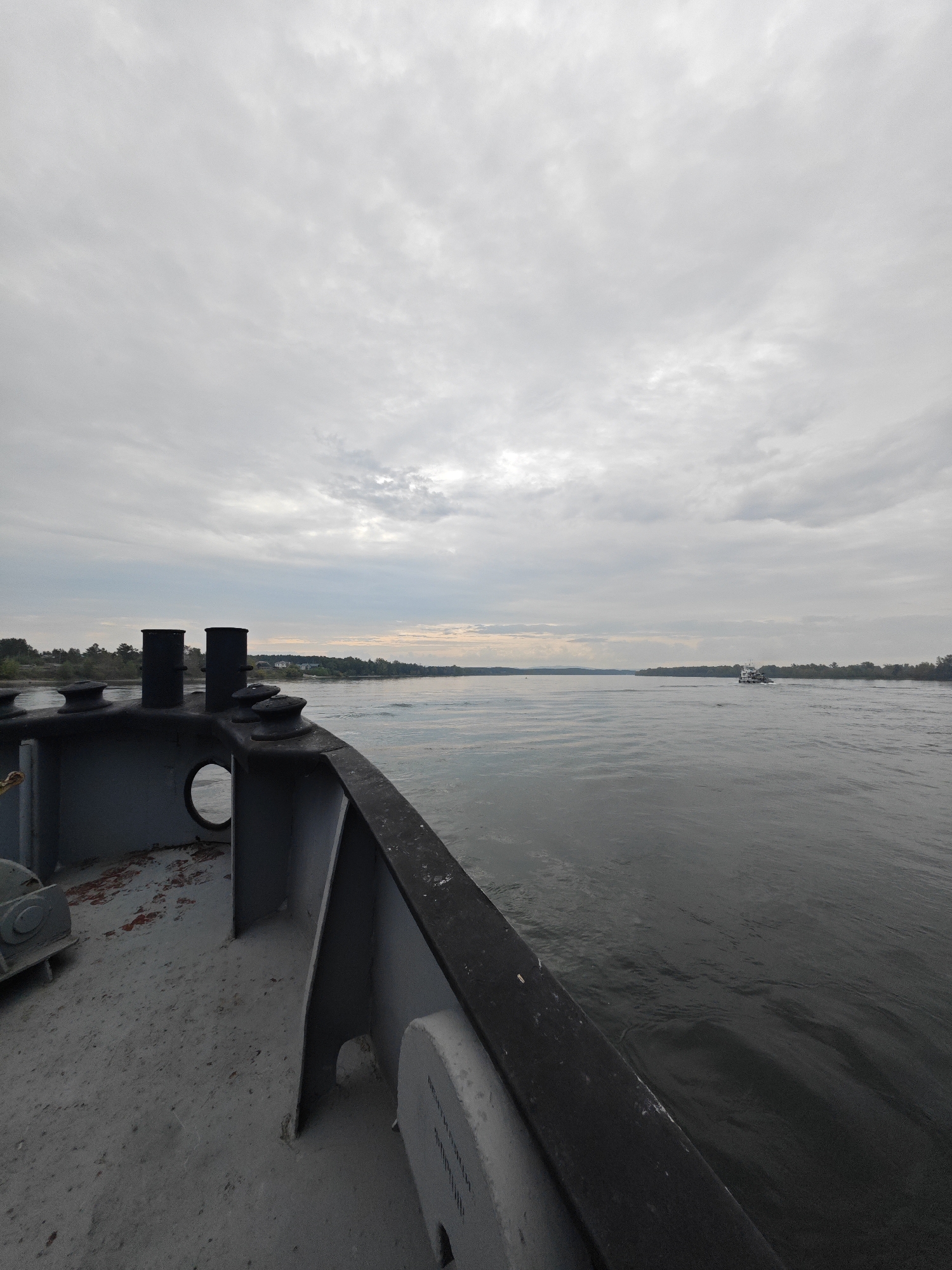 The image depicts a wide, calm river under an overcast sky. In the foreground, the deck of a vessel is visible, showing grey painted metal surfaces and two dark, cylindrical smokestacks. A circular opening is present in the deck structure. In the distance, the river stretches towards a tree-lined horizon, with a small white boat visible in the middle ground. The overall scene is one of a quiet, possibly early morning or late afternoon, journey on the water.