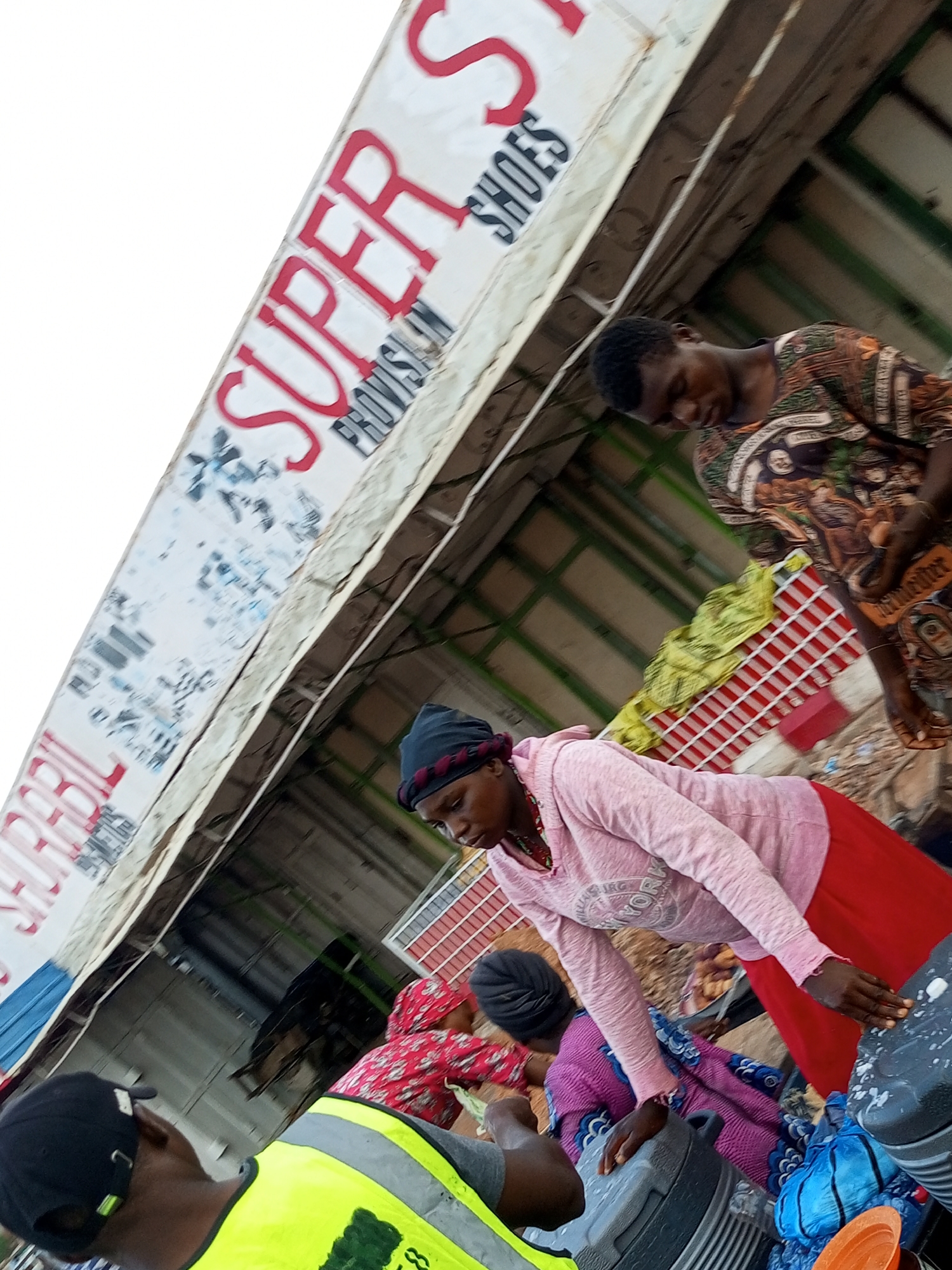 In Tempure, Nigeria, an outdoor market scene unfolds. A large, angled banner with red lettering announcing "SUPER PROVISION SHOES" dominates the background, with some of the text obscured. Beneath the banner, people are engaged in various activities. In the foreground, a person wearing a yellow and grey high-visibility vest and a black cap faces away from the camera, their body angled as if in conversation or observation. To their right, a woman in a pink hoodie and red skirt leans forward, looking down. Behind her, another woman in a purple garment and a black headscarf also looks downwards. Several large, grey containers are visible, some with white splatters. A bright orange object, possibly a lid or plate, rests on one of the containers. The ground appears to be dirt or gravel, with some scattered produce, including yellow items on a red and white checkered rack. The overall atmosphere suggests a bustling marketplace environment.