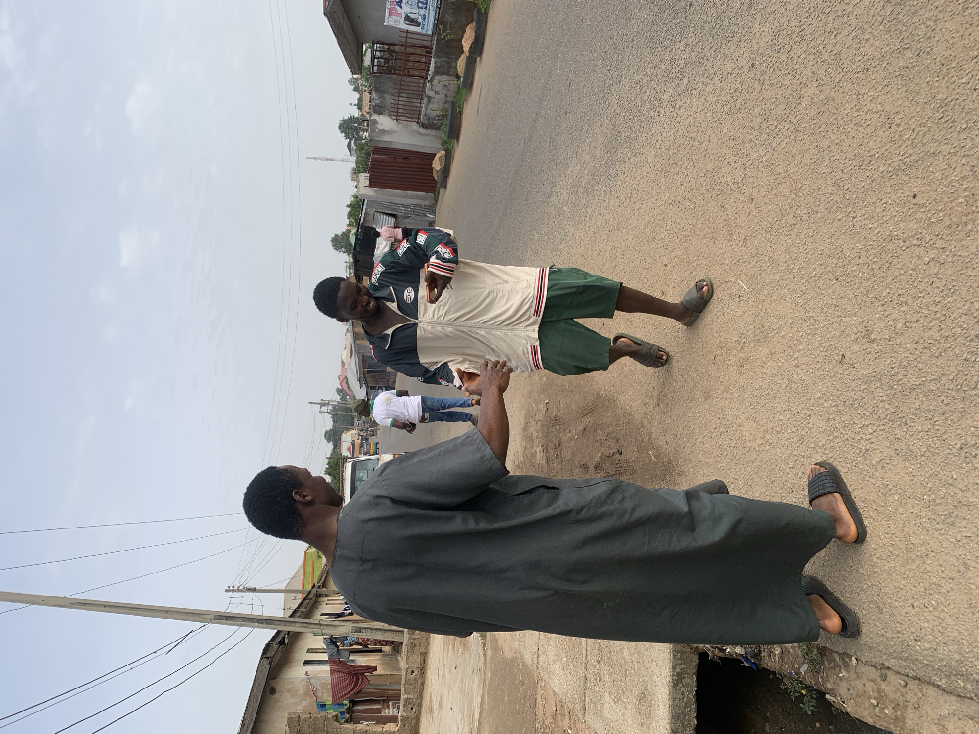 Two men stand on a paved road in Jos, Nigeria. One man wears a dark grey robe and dark sandals. The other wears a black, white, and red jacket, green shorts, and dark sandals. They face each other. In the background, a person in a white shirt and jeans walks away from the camera. Buildings, utility poles, and overhead wires are visible on either side of the road. A political campaign poster is affixed to a building in the background. The sky is overcast.