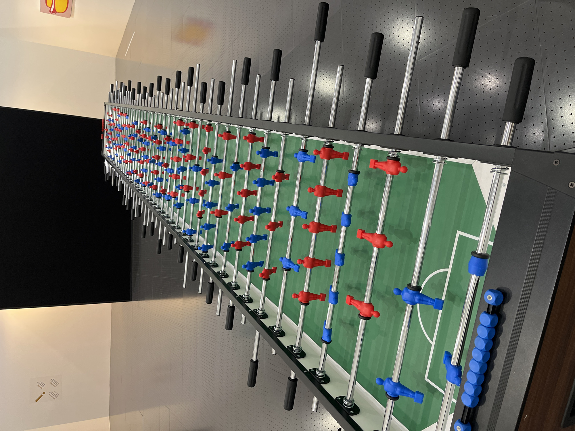 The image shows a foosball table, also known as table football, in Metz, France. The table is set up with opposing teams of red and blue players arranged on metal rods. The playing field is green with white lines marking the boundaries and center. Black handles are attached to the rods for controlling the players. The background includes a wall with a perforated pattern and a black surface to the side. The presence of the foosball table suggests a recreational space, possibly a game room or entertainment area, within Metz.