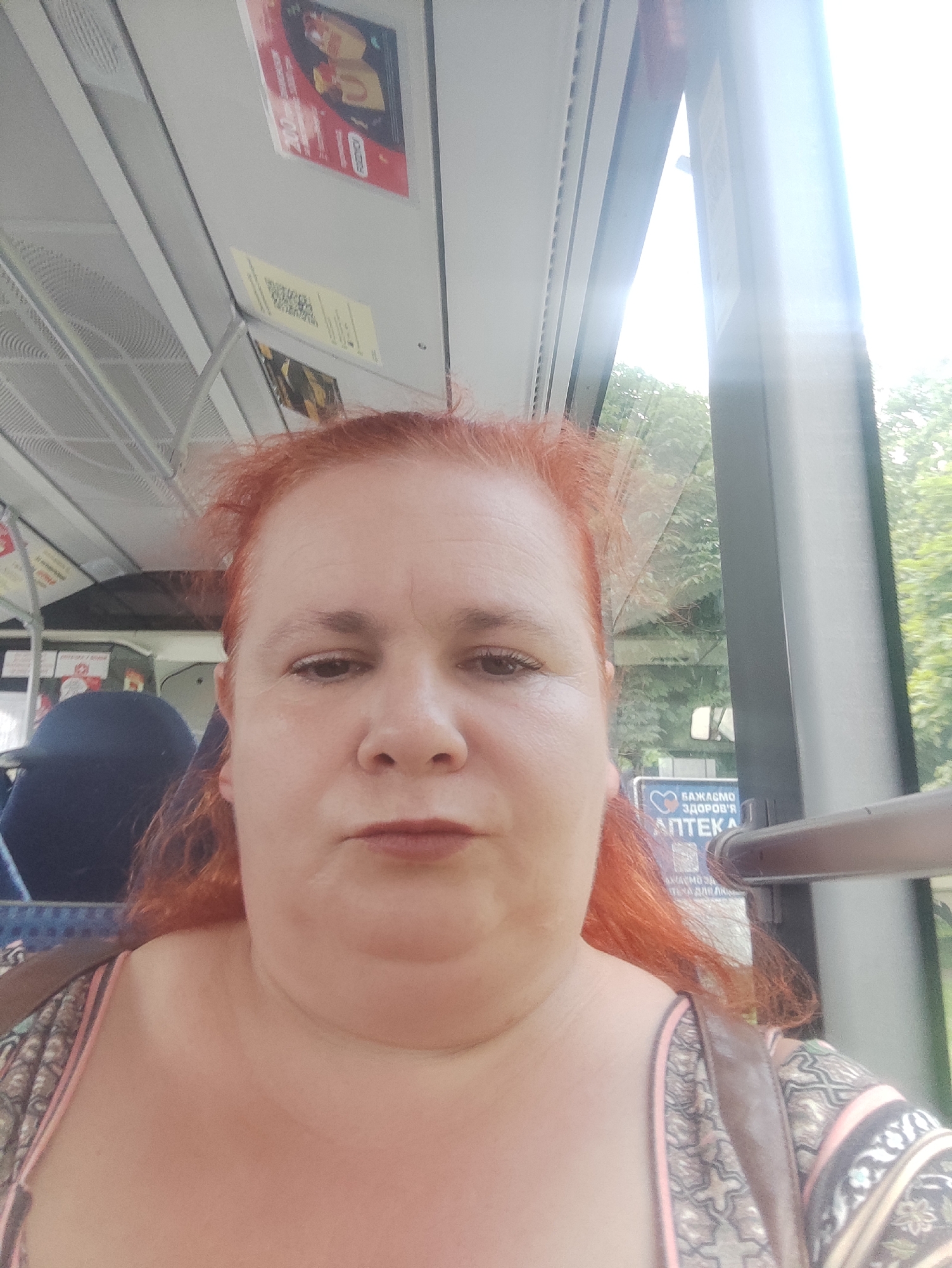 A woman with red hair is seated inside a vehicle, likely a bus or tram, as indicated by the interior ceiling structure and handrails. She is looking directly at the camera, her expression neutral. Visible in the background through the window are trees and greenery, suggesting an outdoor environment. A sign with Cyrillic text, "БАЖАЄМО ЗДОРОВ'Я АПТЕКА," meaning "We wish you health, Pharmacy," is also visible, indicating the location is likely in Ukraine, and specifically Chernivtsi, Ukraine. The lighting suggests it is daytime, with sunlight streaming in from the right.