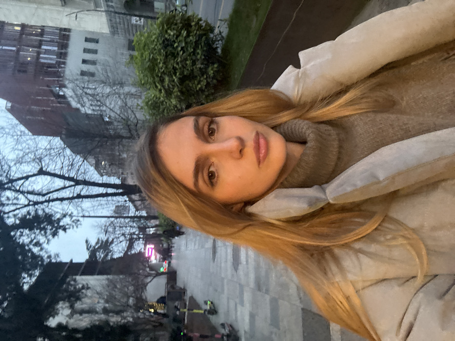 A young adult female is depicted in a close-up, selfie-style outdoor portrait taken at dusk or early evening in the city of Unknown, Unknown. She has light skin, long straight blonde hair, and brown eyes, wearing a light grey puffer jacket over a dark grey turtleneck sweater, looking directly at the camera with a neutral expression. The background reveals an urban environment with a paved sidewalk stretching into the distance on the left. Several green and black electric scooters are parked along the sidewalk. Beyond the sidewalk, a road is visible, bordered by green bushes. In the midground and background, multiple buildings of varying heights and architectural styles are present, including one with a distinctive reddish-brown facade. Bare deciduous trees and evergreen foliage are distributed throughout the scene. Distant figures walk along the sidewalk, and ambient artificial lights are visible. The sky is overcast and grey-blue.
