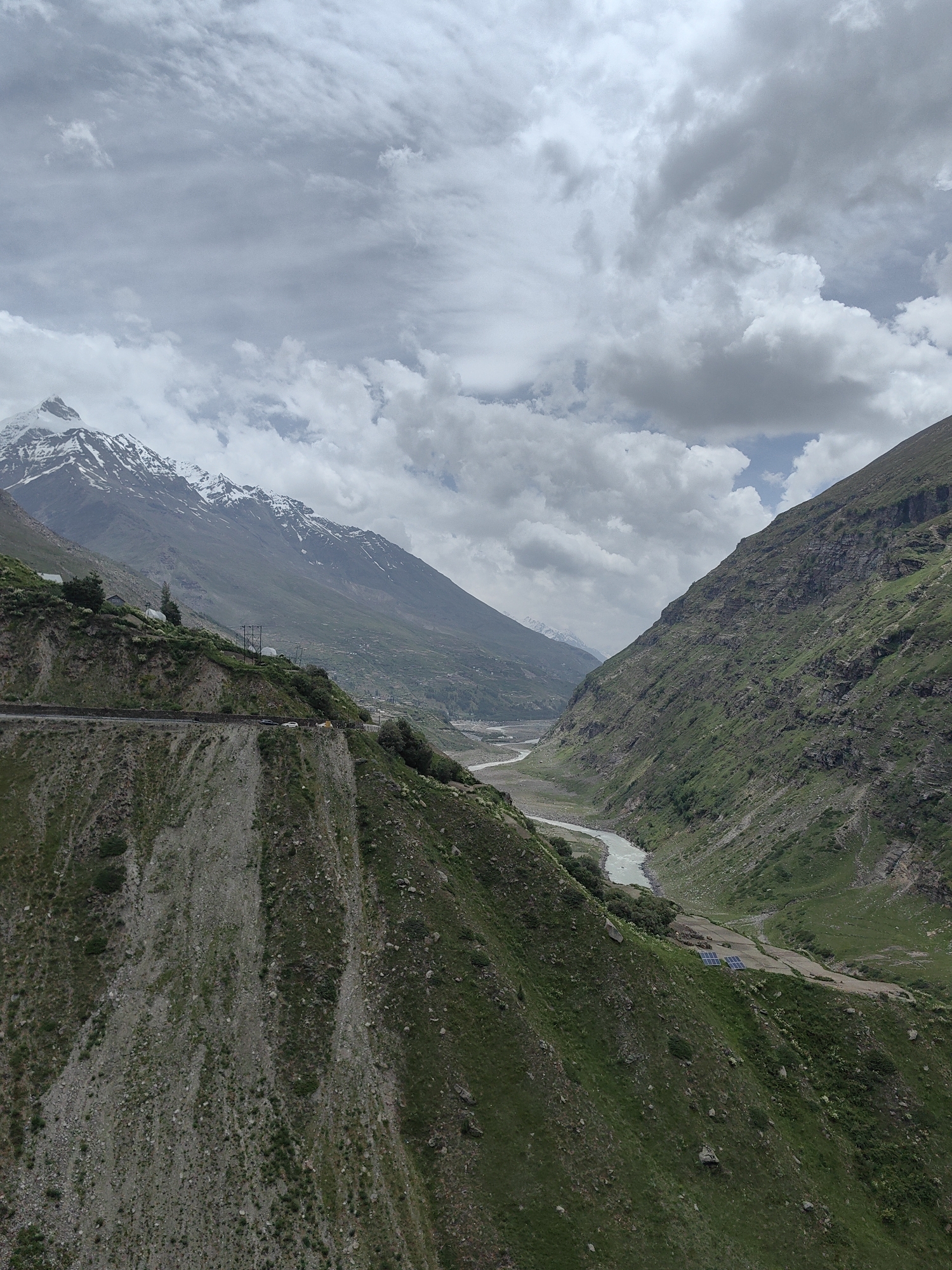 The media file captures a panoramic view of a mountainous landscape in Yamling, India. The scene is dominated by steep, green, and rocky slopes, with a winding river flowing through the valley below. In the foreground, a dirt road snakes along the side of a precipice, with a few vehicles visible, suggesting ongoing travel or transport. The mountains in the distance are capped with snow, indicating a high-altitude region. The sky is overcast with dramatic, heavy clouds, hinting at potentially turbulent weather or a recent storm. The overall mood is one of rugged natural beauty and a sense of remoteness. The presence of solar panels on a small plateau suggests some human habitation or infrastructure in this otherwise wild setting. There is no visible text in the media file.