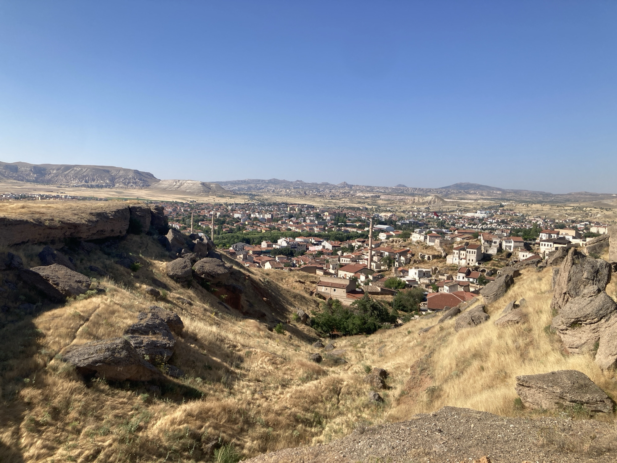 This is an eye-level, wide-angle photograph of a valley with a town nestled in the distance, taken on a clear, sunny day. The foreground is dominated by a steep, dry, grassy slope punctuated by large, dark, weathered rocks. The dry grass is golden-yellow and appears wind-swept. The terrain descends into a valley where the town of Avanos İlçe Merkezi, Turkey, is visible. The town features a cluster of buildings with red-tiled roofs and white or light-colored walls, characteristic of the region. Several minarets rise above the rooftops, indicating the presence of mosques. Beyond the town, the landscape opens up to rolling hills and mountains under a clear blue sky, with no clouds present. The overall impression is one of a dry, arid environment with a historic settlement.