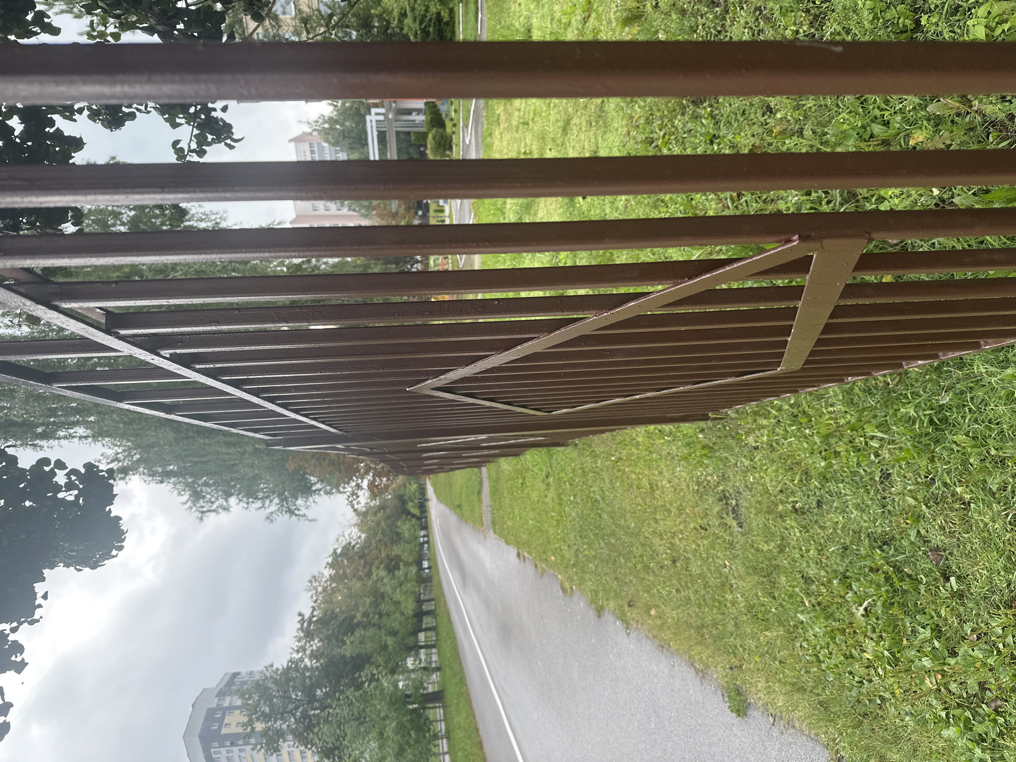 A vertical, medium shot shows a brown metal fence in the foreground, with its vertical bars and a diamond-shaped decorative element on the right side. Beyond the fence, a wet asphalt road with a white line curves to the left, bordered by green grass. In the background, trees line the road, and apartment buildings are visible. The sky is overcast and cloudy, suggesting a damp or rainy day. The location is identified as Minsk, Belarus. There are no people visible in the image.