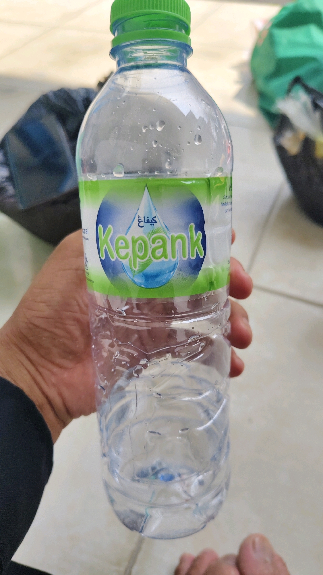 A person's hand holds a partially full bottle of Kepank mineral water. The bottle is made of clear plastic with a green cap. The label on the bottle is predominantly green and blue, featuring a stylized water droplet and the brand name "Kepank" in large green letters. The bottle is condensation-covered. In the background, slightly out of focus, are a dark blue bag and a green plastic bag. The scene appears to be outdoors, on a light-colored tiled surface. The lighting suggests daytime.