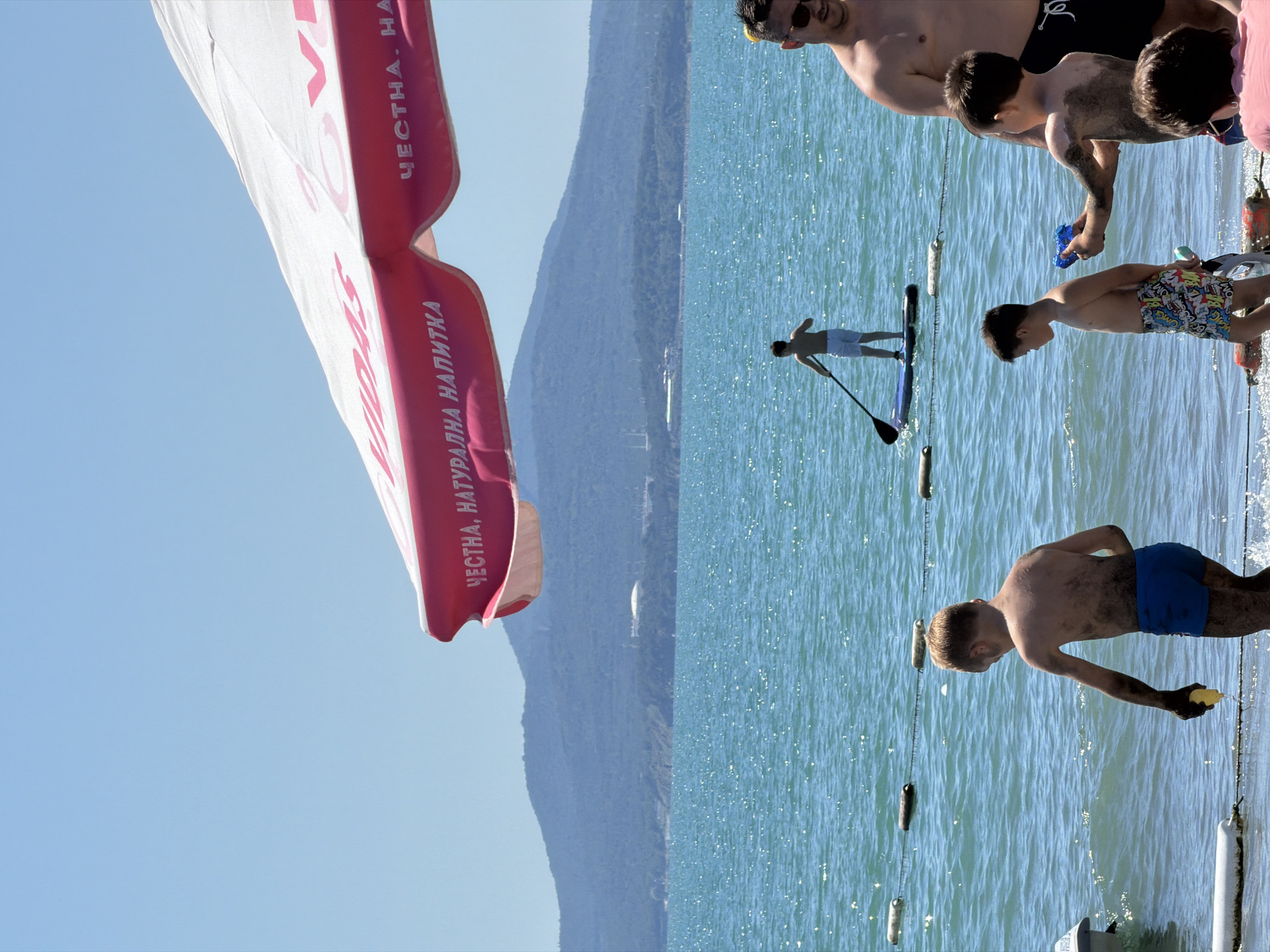 The image depicts a sunny day at a beach, likely in Burgas, Bulgaria, as indicated by the presence of a large umbrella with Cyrillic text that translates to "Honest, Natural Drink." The sea is a bright turquoise, with small waves and reflections of the sun. In the foreground, three young boys are on the shore. One boy, wearing blue swim trunks, is covered in mud and holds a yellow object in his hand. Another boy, in patterned swim trunks, stands next to him. A third boy, partially obscured by the others, appears to be holding a blue water gun. To the right, a man with sunglasses and a bare chest looks towards the sea. Further out in the water, a lone figure stands on a paddleboard, holding a paddle. In the background, a chain of buoys stretches across the water, and a distant, tree-covered mountain range with some buildings is visible under a clear blue sky.