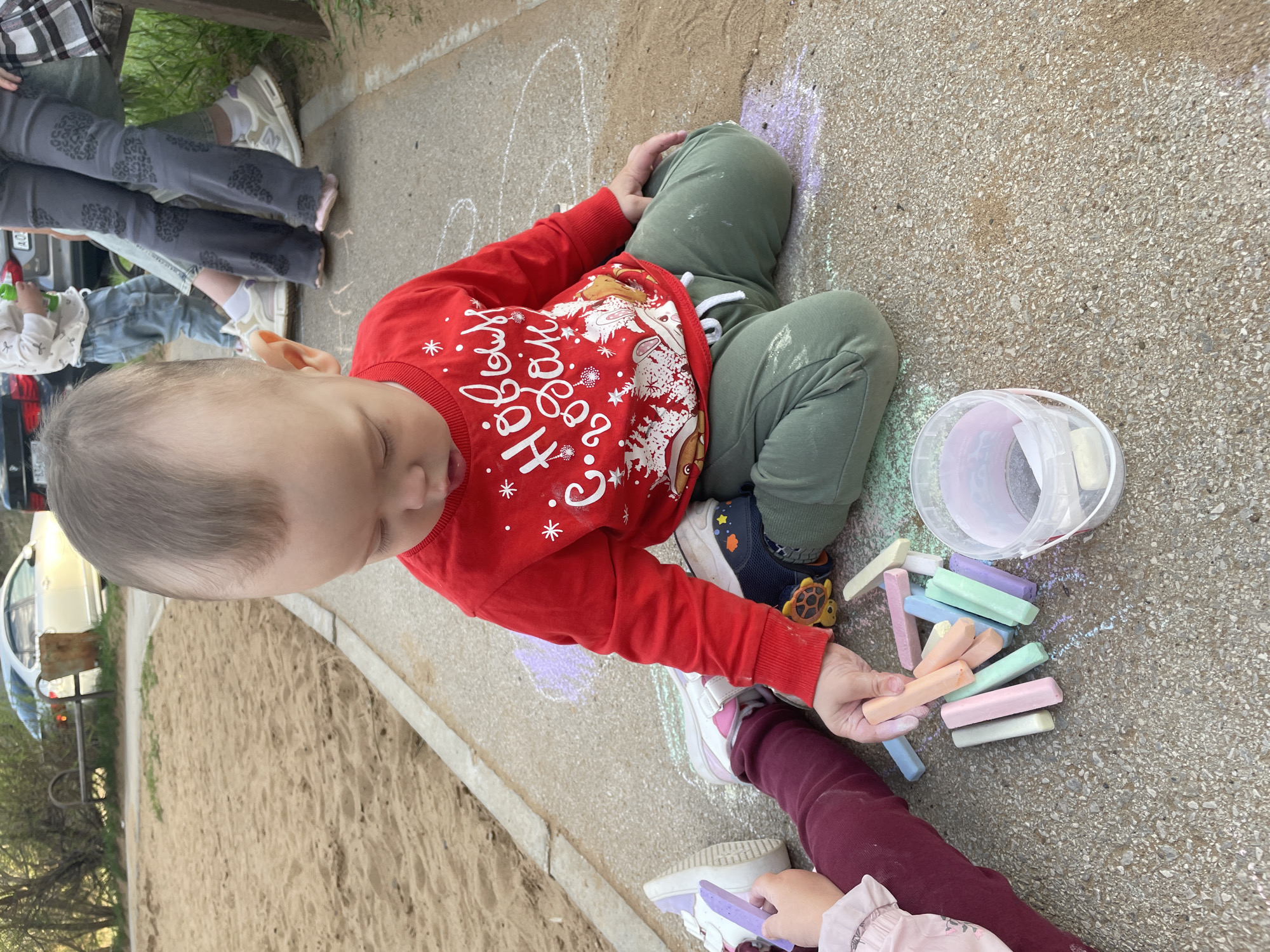 A child wearing a red Christmas-themed sweater and green pants sits on a concrete surface, surrounded by colorful sidewalk chalk. Next to the child is a bucket of chalk. Another person, wearing burgundy leggings and white shoes, is also holding a piece of chalk. The location appears to be outdoors, next to a sandbox area. Based on visual cues and image content, it is not possible to confirm the location in the city of Perm, Russia.