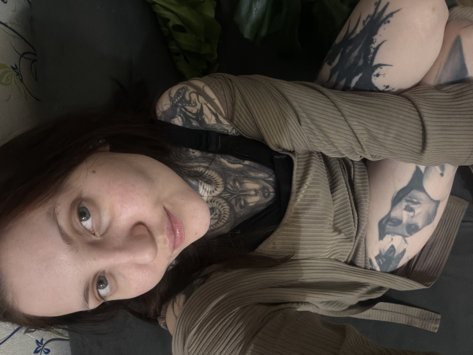 A woman with brown hair, pale skin, and distinctive tattoos lies on a dark-colored surface, possibly a couch or bed. She wears a light brown, ribbed sweater or cardigan. Visible tattoos adorn her neck, arms, and thighs, depicting faces and abstract designs. A houseplant with large green leaves is positioned behind her. Her face is oriented towards the camera, with a direct gaze and a subtle smile.
