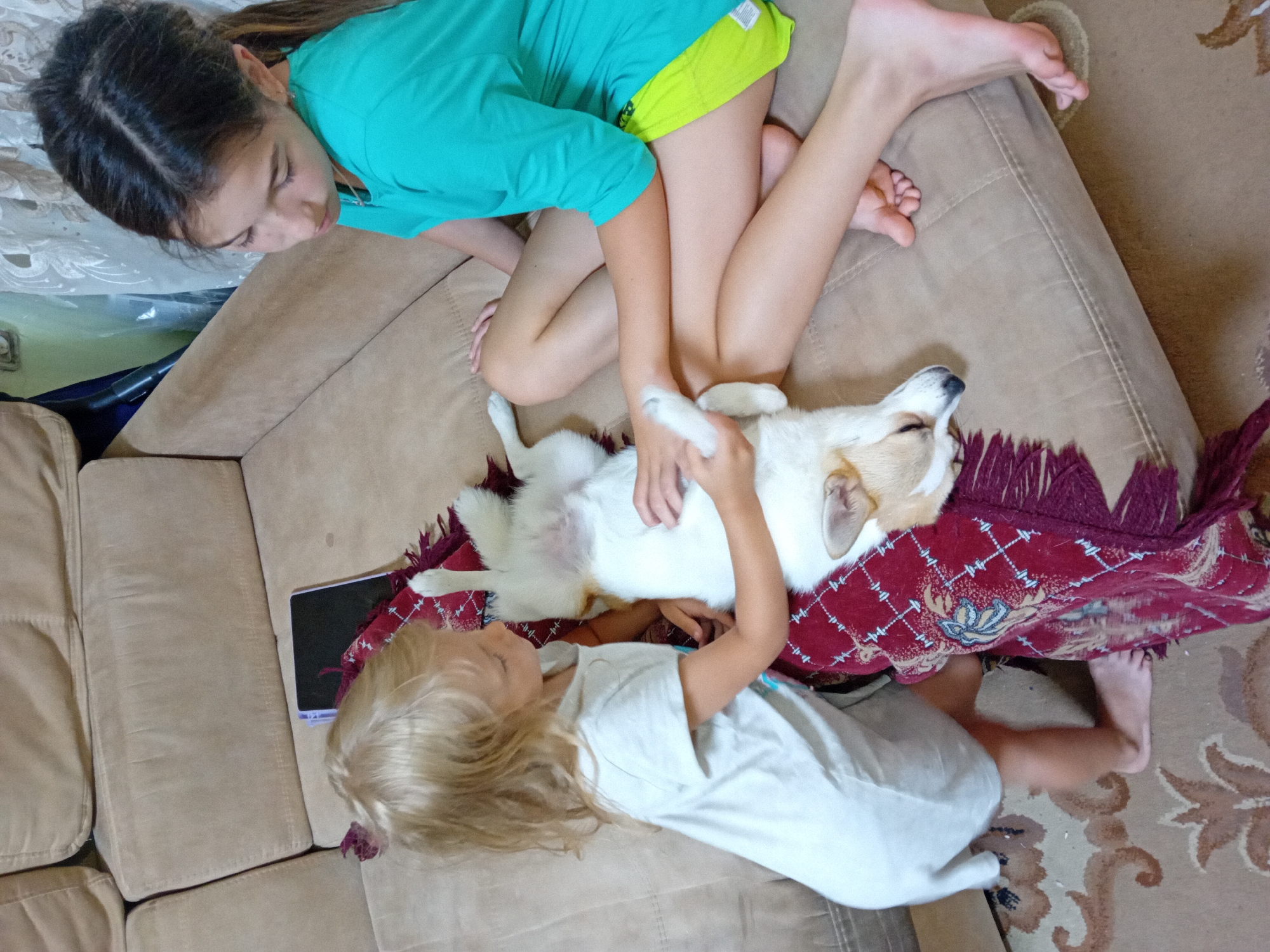 Two young girls are interacting with a corgi dog on a beige sofa. The girl on the left, with blonde hair, is wearing a white t-shirt and shorts, and is petting the dog. The girl on the right, with dark hair, is wearing a teal t-shirt and lime green shorts, and is also interacting with the dog. The corgi is lying on its back on a patterned blanket. The scene appears to be indoors, possibly in a living room, with a rug visible on the floor. The environment suggests a casual, domestic setting. There is no indication of the specific location within Chuhuiv, Ukraine.