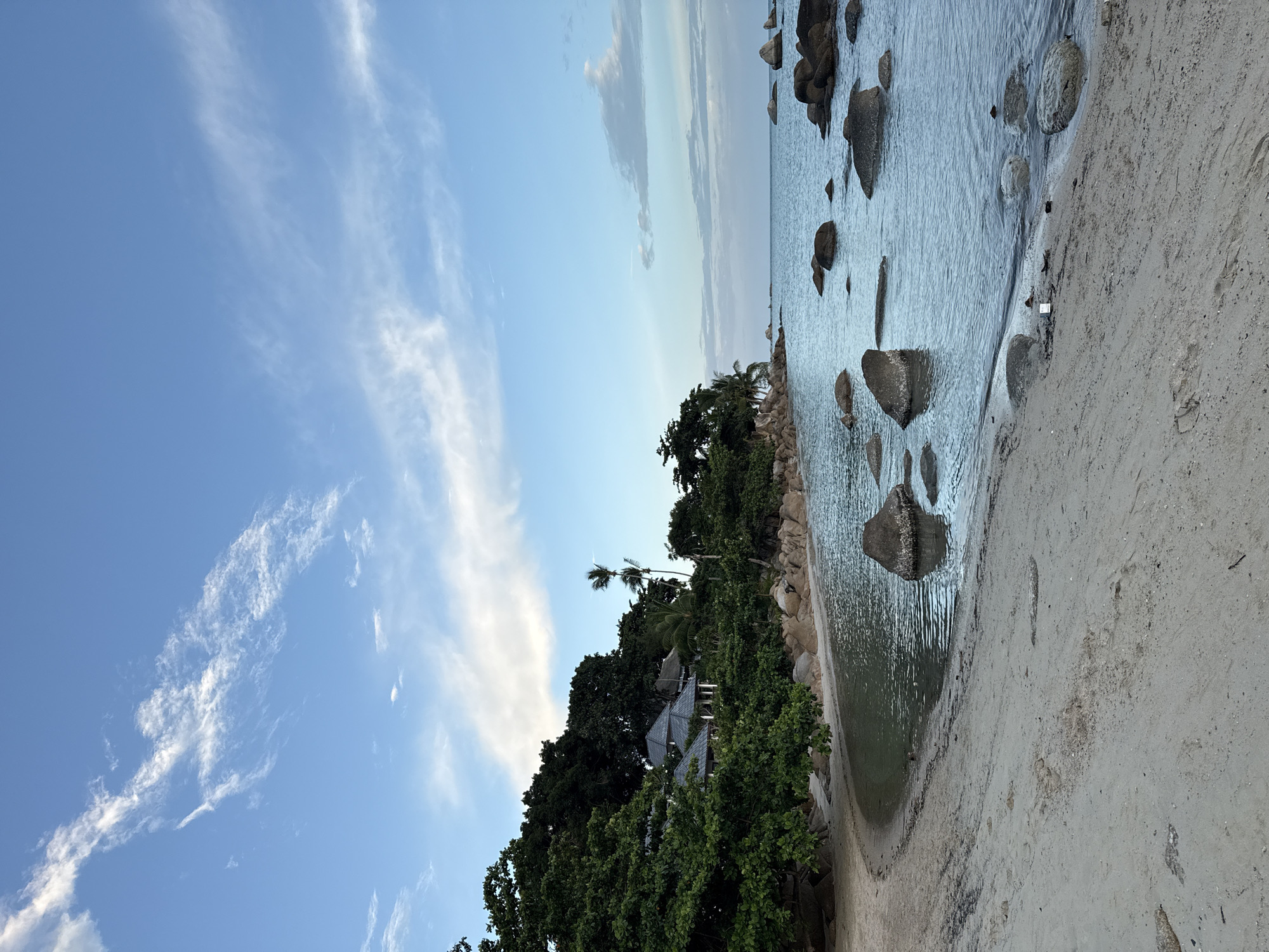 This image depicts a serene tropical beach scene, likely in the Ko Samui District of Thailand. The foreground is dominated by a sandy beach with faint footprints and scattered debris. The calm, blue ocean stretches out towards the horizon, dotted with numerous large, dark rocks submerged in the water. Along the left side of the image, lush green trees and vegetation line the shore, partially obscuring some buildings with grey, sloped roofs, suggesting a resort or residential area nestled within the foliage. The sky is a clear blue with wispy white clouds, indicating fair weather. The overall atmosphere is peaceful and natural.