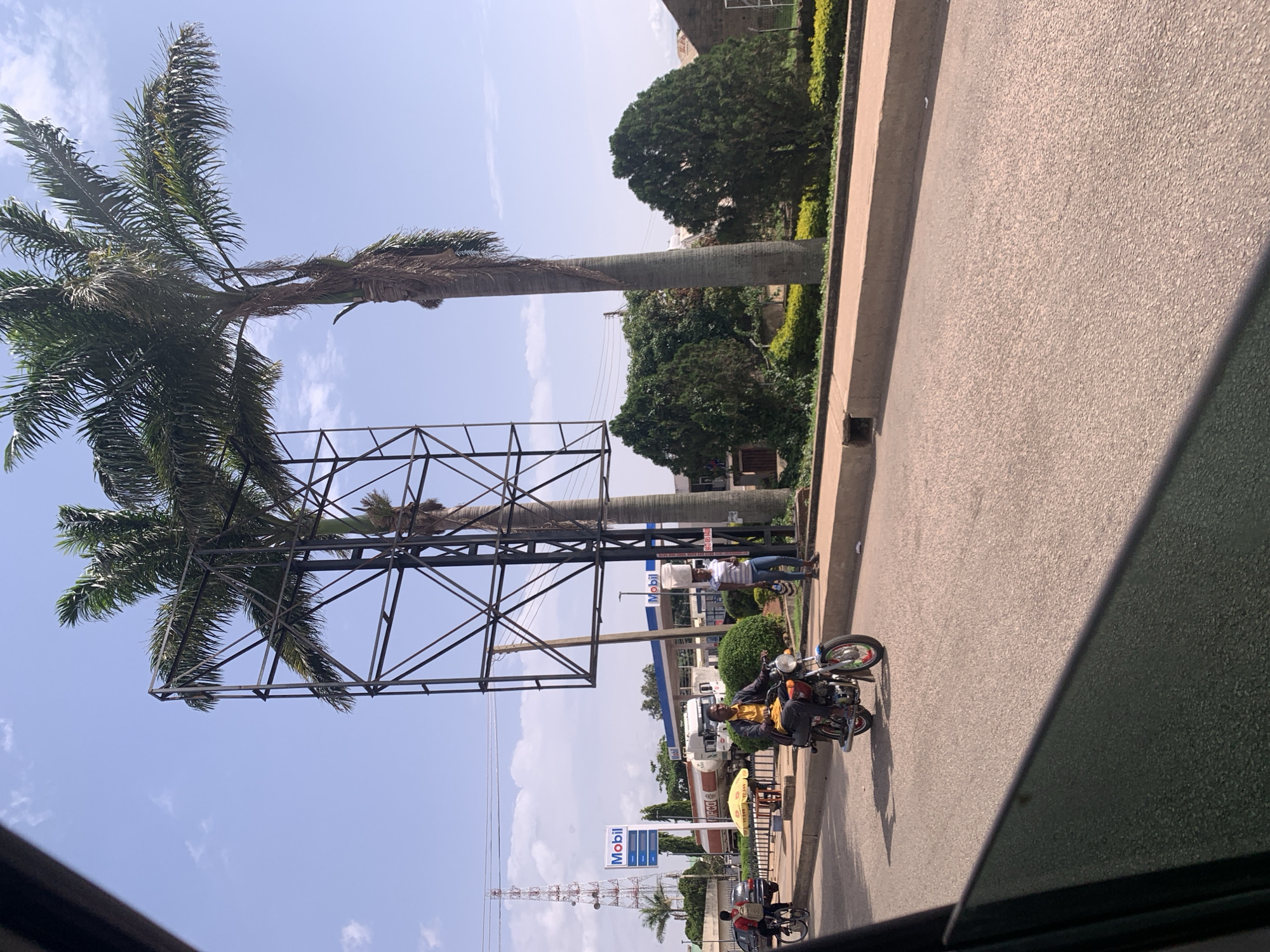The image, captured in Jos, Nigeria, depicts an outdoor scene with a clear blue sky and scattered clouds, suggesting a sunny day. The foreground is dominated by a tall palm tree with a large metal billboard structure adjacent to it. In the middle ground, a man is riding a motorcycle, wearing a yellow shirt and dark pants, casting a shadow on the asphalt road. Further back, a Mobil gas station is visible with its blue and white signage. Two people are standing near the gas station. Behind the gas station, there are several trees and some buildings. The overall impression is a typical roadside view in a warm climate. The image has a bright and slightly overexposed quality, typical of outdoor photos taken in strong sunlight.