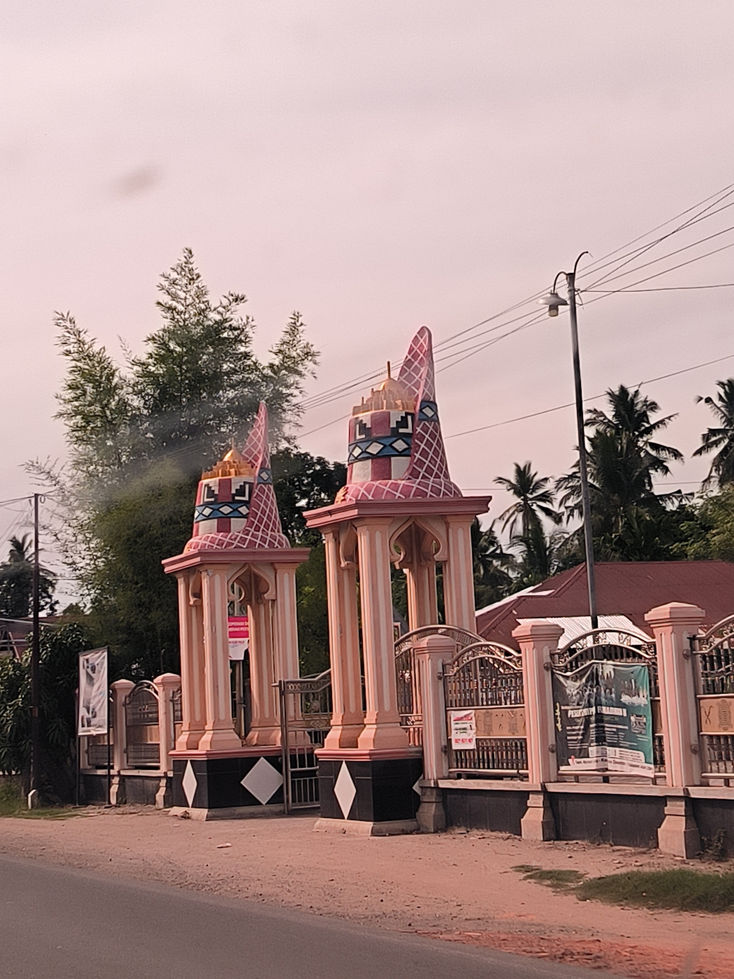 The image shows an ornamental gate, likely leading to a religious or community center, in Palohlada, Indonesia. The gate is flanked by two pink and white pillars topped with conical structures decorated with a diamond pattern and what appears to be a stylized face. Behind the pillars, a metal fence with decorative elements stretches across the frame. A red-tiled roof building is visible beyond the fence. Palm trees and other foliage are present in the background, suggesting a tropical environment. Several banners or posters are attached to the fence, one of which is green and features text and images of people. A lamppost with wires stretches across the upper right portion of the image. The sky is overcast and a pale pinkish hue. The foreground consists of a dirt road and a bit of dry grass. There are no people visibly present in the image.