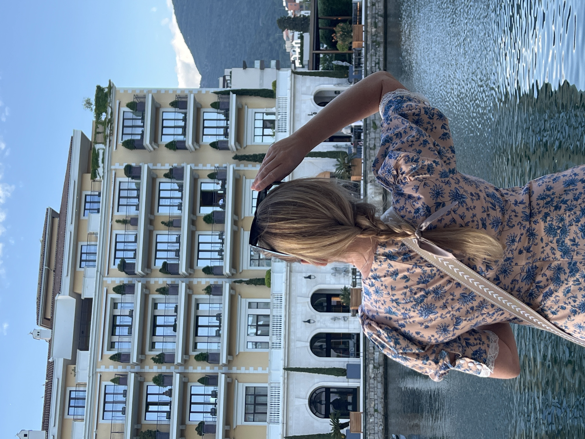 A woman stands near a body of water, facing a tall, light yellow building with multiple balconies, each adorned with plants. The woman has blonde hair in a braid and wears a floral dress. She holds her hand to her forehead, shielding her eyes. Sunglasses are perched on her head. A purse strap is visible over her shoulder. The background includes mountains and a blue sky. The setting appears to be Porto Montenegro in Tivat.