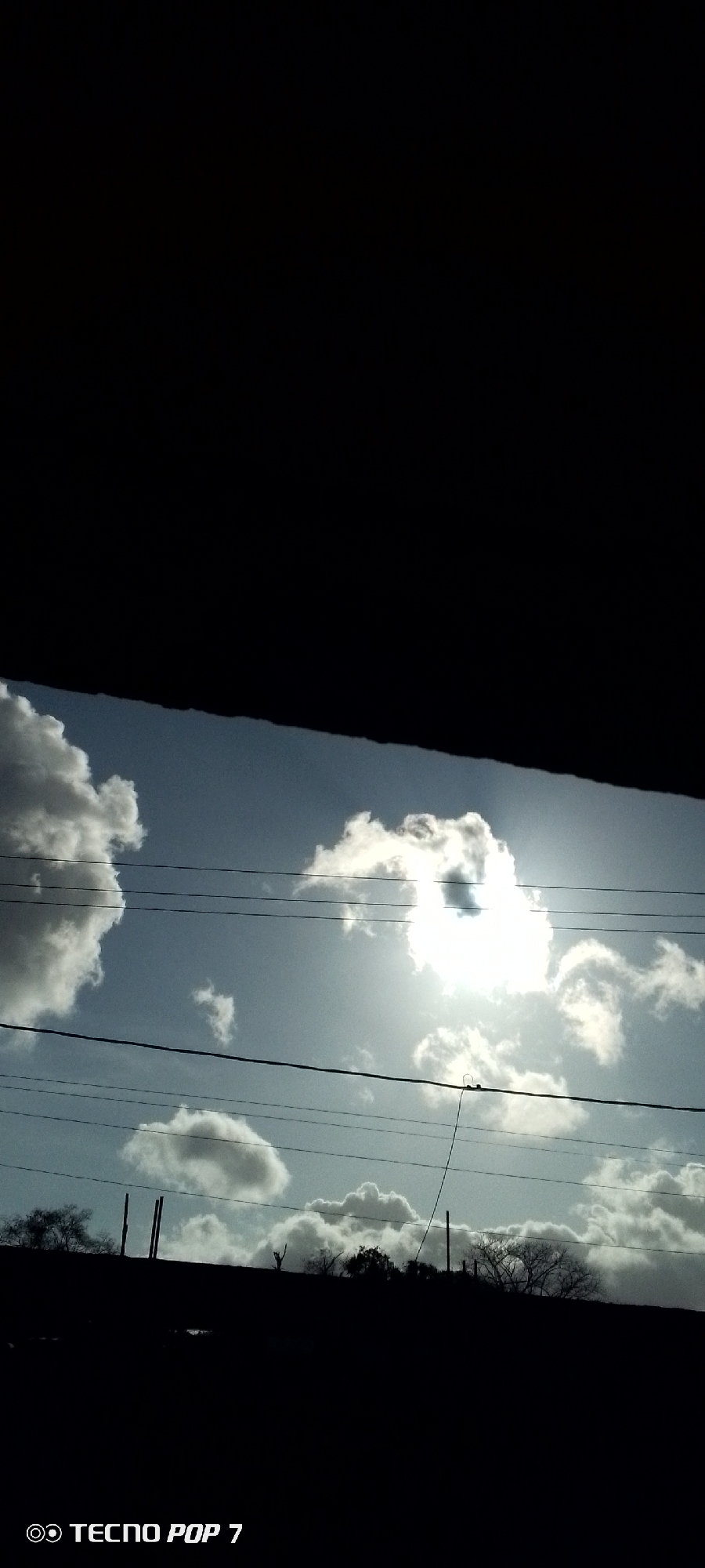 The image shows a view of the sky with clouds and the sun shining through them, partially obscured by dark shadows. There are also utility poles and wires visible against the sky. The ground or a structure is silhouetted at the bottom of the frame, along with trees and shrubs in the landscape. The lower left corner of the picture has the “TECNO POP 7” text marking. The location in the city of Altagracia, Venezuela, cannot be verified from the visual content alone. No people are visible in the image.