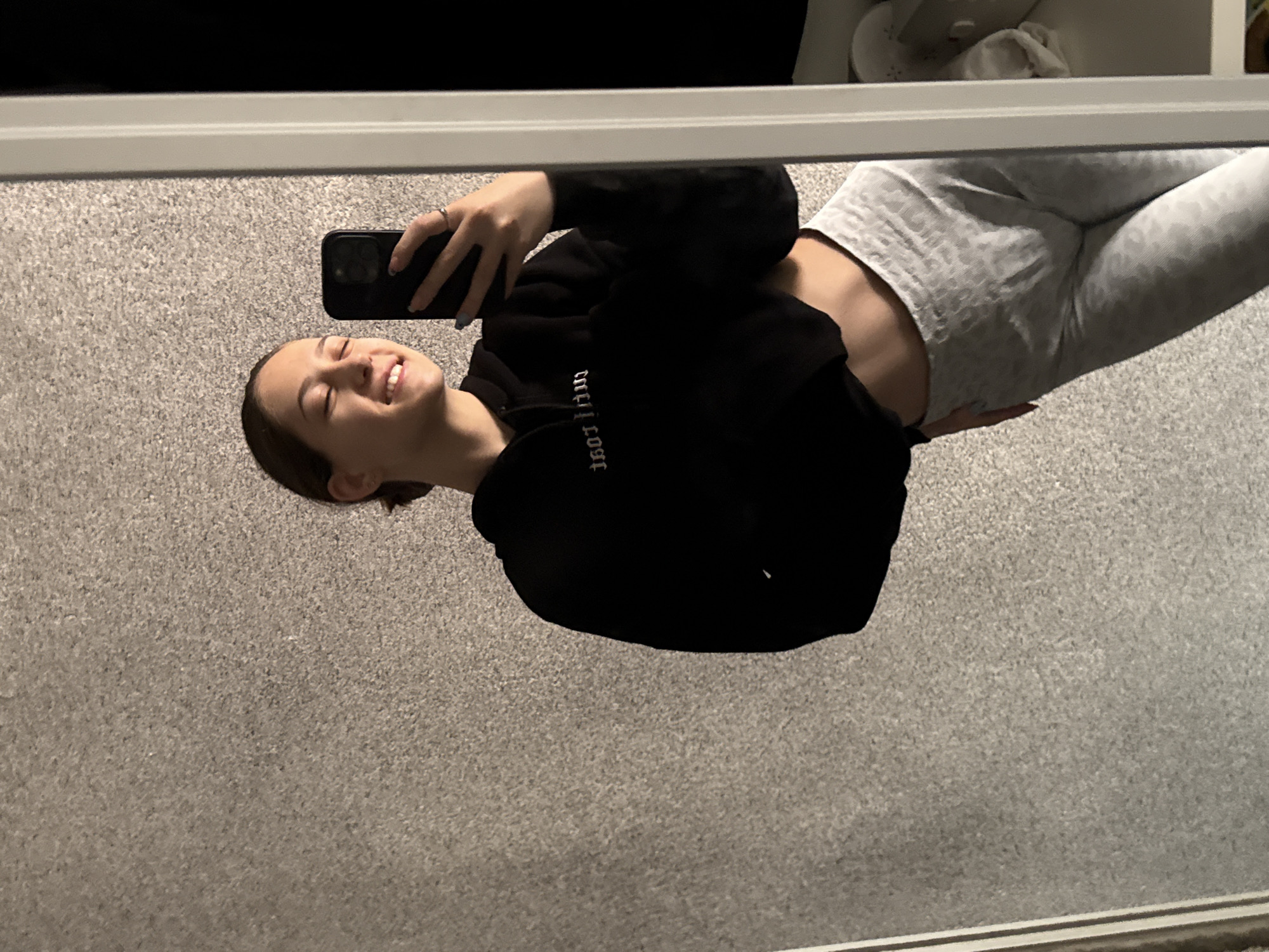 A young woman is reflected in a mirror, taking a selfie. She is wearing a black hoodie and grey patterned leggings, exposing her midriff. She's holding a black phone and smiling, facing upwards. The background appears to be a gray carpet and a piece of furniture above. There is no evidence in the image to indicate that it was taken in Sofia, Bulgaria.