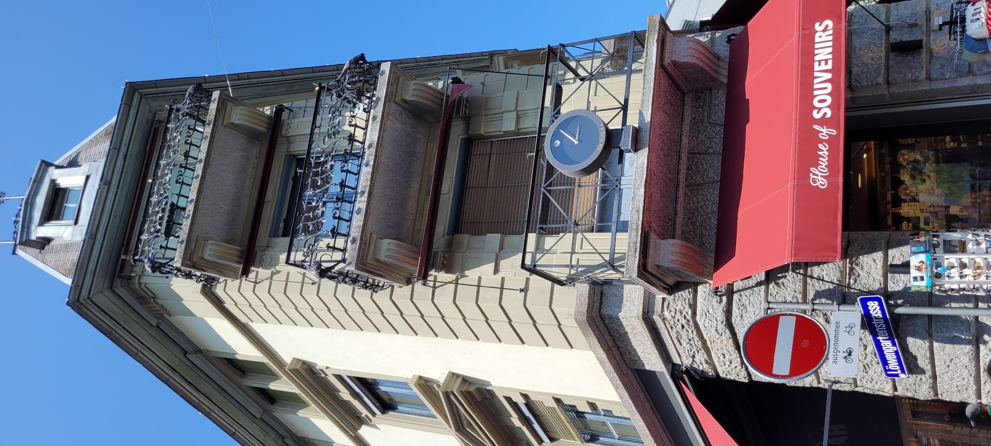 The image shows a street view in Wattens, Austria, focusing on the facade of a building. The building has a cream-colored exterior with stone detailing around the base and corners. It features multiple balconies with ornate black metal railings. A prominent dark clock with a white face and hands, displaying the time as approximately 12:08, is mounted on the railing of a mid-level balcony. The building has red awnings over storefronts at street level. One awning displays the text "House of SOUVENIRS" in white script. Below the awnings, there are signs, including a red circular "no entry" sign and a blue street sign that reads "Löwengartenstrasse". The sky above is clear and blue. The scene is captured from a low-angle perspective, emphasizing the height of the building. There are no people clearly visible or identifiable in the image.