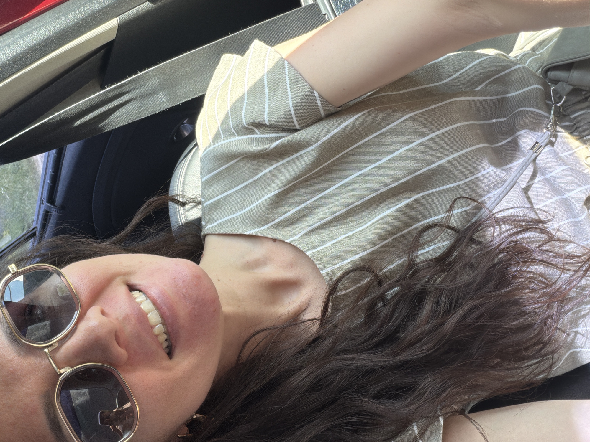 A close-up, over-the-shoulder shot captures a woman with long, dark, wavy hair, wearing sunglasses and a light-colored, striped blouse. She is seated in a car, as indicated by the presence of a seatbelt and car interior visible in the background. The lighting suggests it is daytime, with sunlight casting shadows on the seatbelt. The woman is smiling, and her face is partially obscured by her sunglasses. Her phone is visible reflected in her sunglasses, showing a selfie.