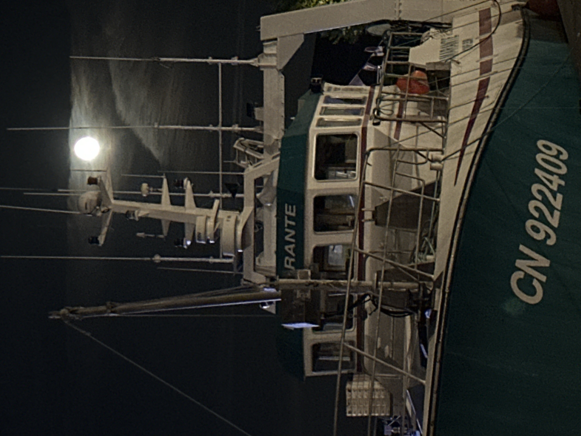 The image captures a fishing trawler docked at night in the port of Honfleur, France. The moon is visible in the dark sky, partially obscured by wispy clouds. The boat, painted predominantly teal and white, has the registration "CN 922409" clearly marked on its hull. The name "AMARANTE" is also visible on the stern. The vessel features a multi-level superstructure with windows and various antennae and radar equipment. The deck area shows railings, crates, and a life preserver. The overall scene suggests a quiet night in the harbor.