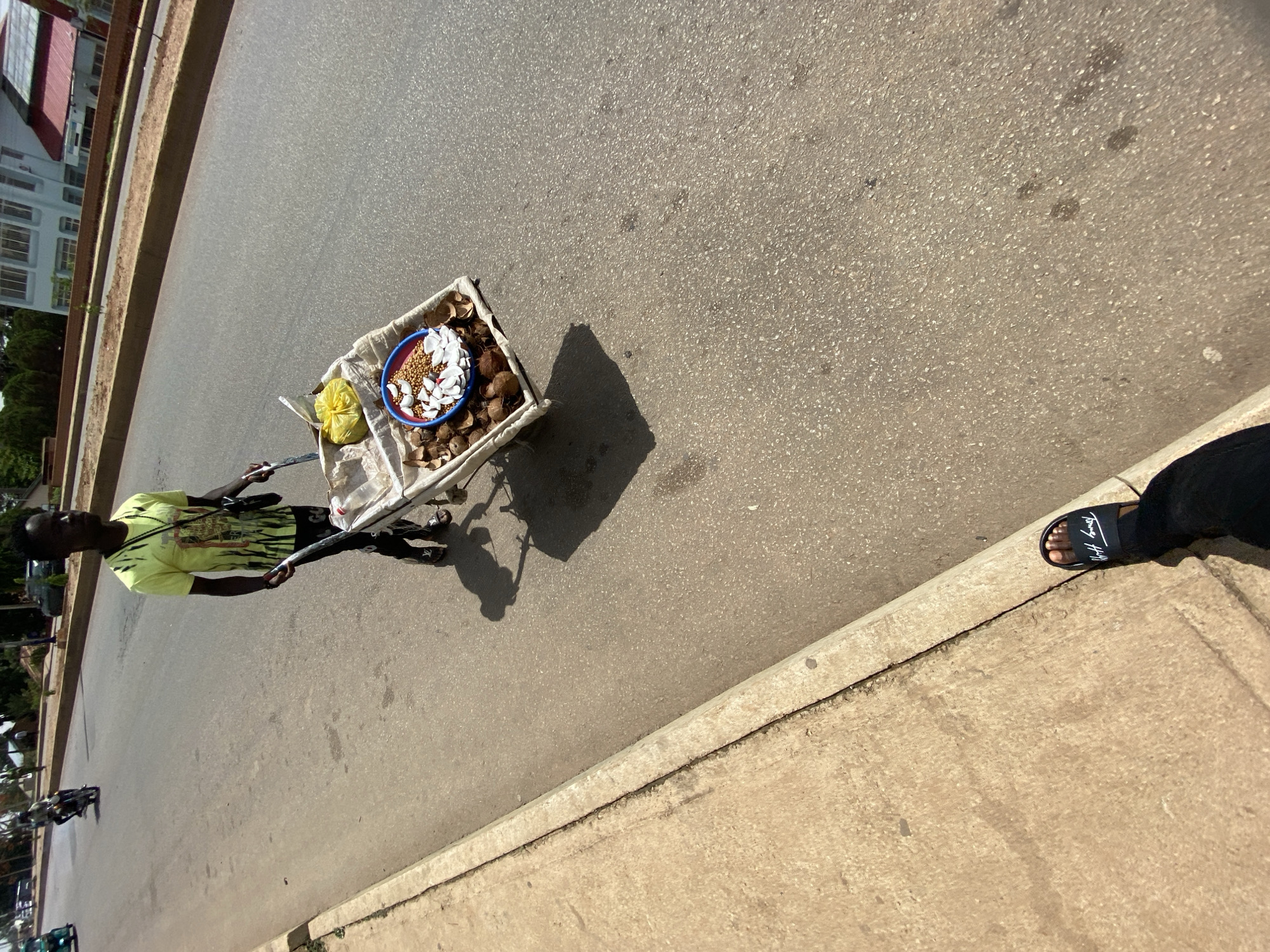 An aerial shot reveals a street scene in Bukuru, Nigeria. A man in a chartreuse t-shirt pulls a wheeled cart. The cart contains coconuts, coconut shavings, and what appears to be a small bag. The man faces left, pulling the cart away from the viewer. The street is paved and flanked by sidewalks. In the lower right corner, a foot wearing a black sandal is visible. A multistory building is partially visible on the left side of the frame.