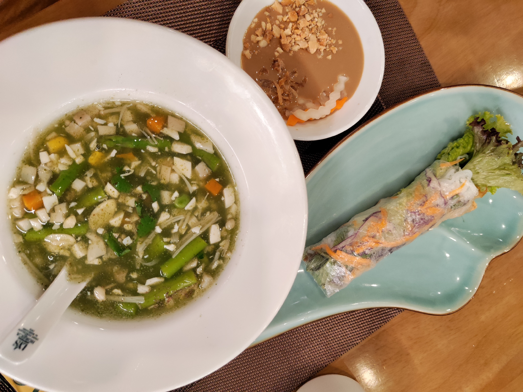 The image shows a meal consisting of a white bowl of green soup with tofu, asparagus, and carrots, a white bowl of peanut sauce with crushed peanuts and pickled vegetables, and a spring roll on a light blue plate. These items are arranged on a brown woven placemat on a wooden table. The photo may be taken in Da Lat, Vietnam, based on the style of the meal.