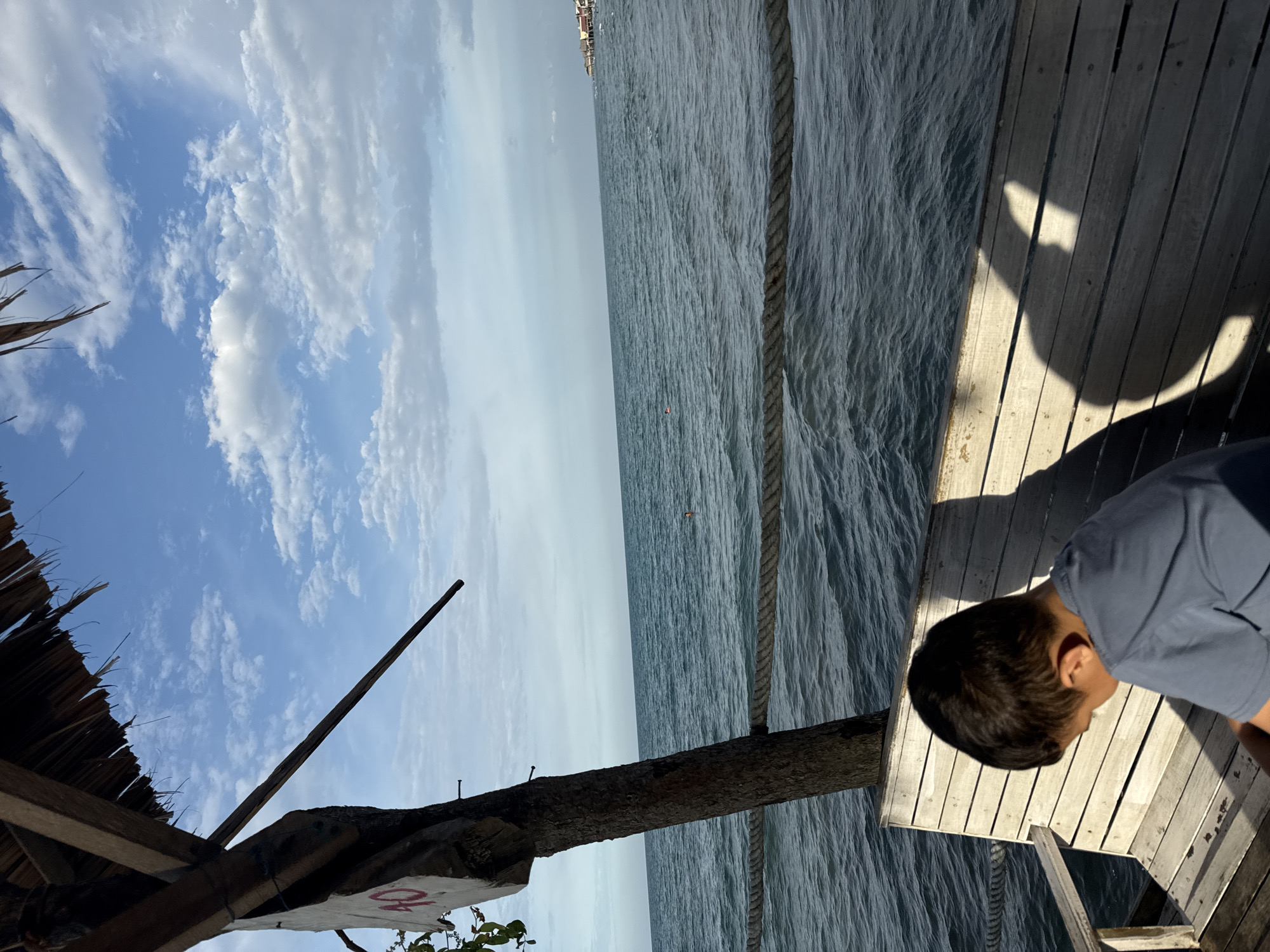 An overhead view from a wooden structure looking out at the ocean. In the foreground, a young boy with dark hair, wearing a blue t-shirt, sits on a weathered wooden bench, his head bowed. His shadow is cast onto the wooden planks of the bench. To the left, a rough-hewn wooden post, part of the structure, has a white sign with a red numeral "10" affixed to it. A thick, braided rope is strung horizontally across the frame, separating the foreground from the ocean. The ocean is a deep blue-green, with gentle waves. In the distance, a small pink buoy is visible on the water, and further out, a cluster of buildings with red roofs can be seen along the coastline, suggesting this is likely a beach resort area. The sky above is a bright blue, dotted with scattered white clouds. The scene appears to be in Lhokseumawe, Indonesia, likely a seaside location.