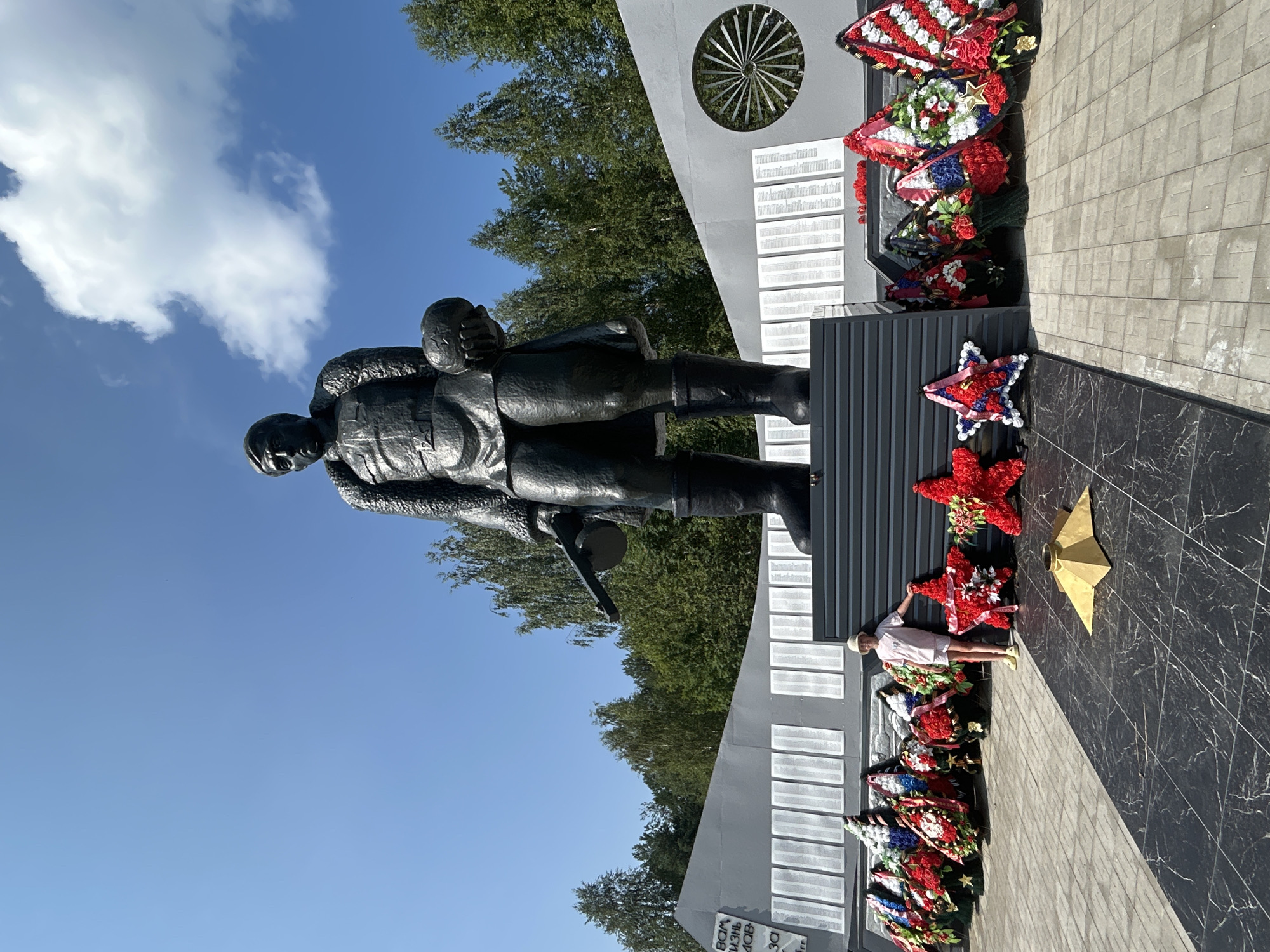 A low-angle, medium shot captures a monumental black statue of a soldier holding a globe in a memorial park on a sunny day. The statue stands on a grey platform, with a large grey wall behind it, inscribed with lists of names. In front of the statue are several large, colorful wreaths and two red, star-shaped floral arrangements, one of which a young girl in a light-colored outfit and hat is standing next to. The ground in the foreground is paved with black and grey tiles, and in the distance, a line of trees forms a backdrop against a clear blue sky with scattered white clouds. This is the monument to the fallen soldiers in the city of Krasnovishersk, Russia.