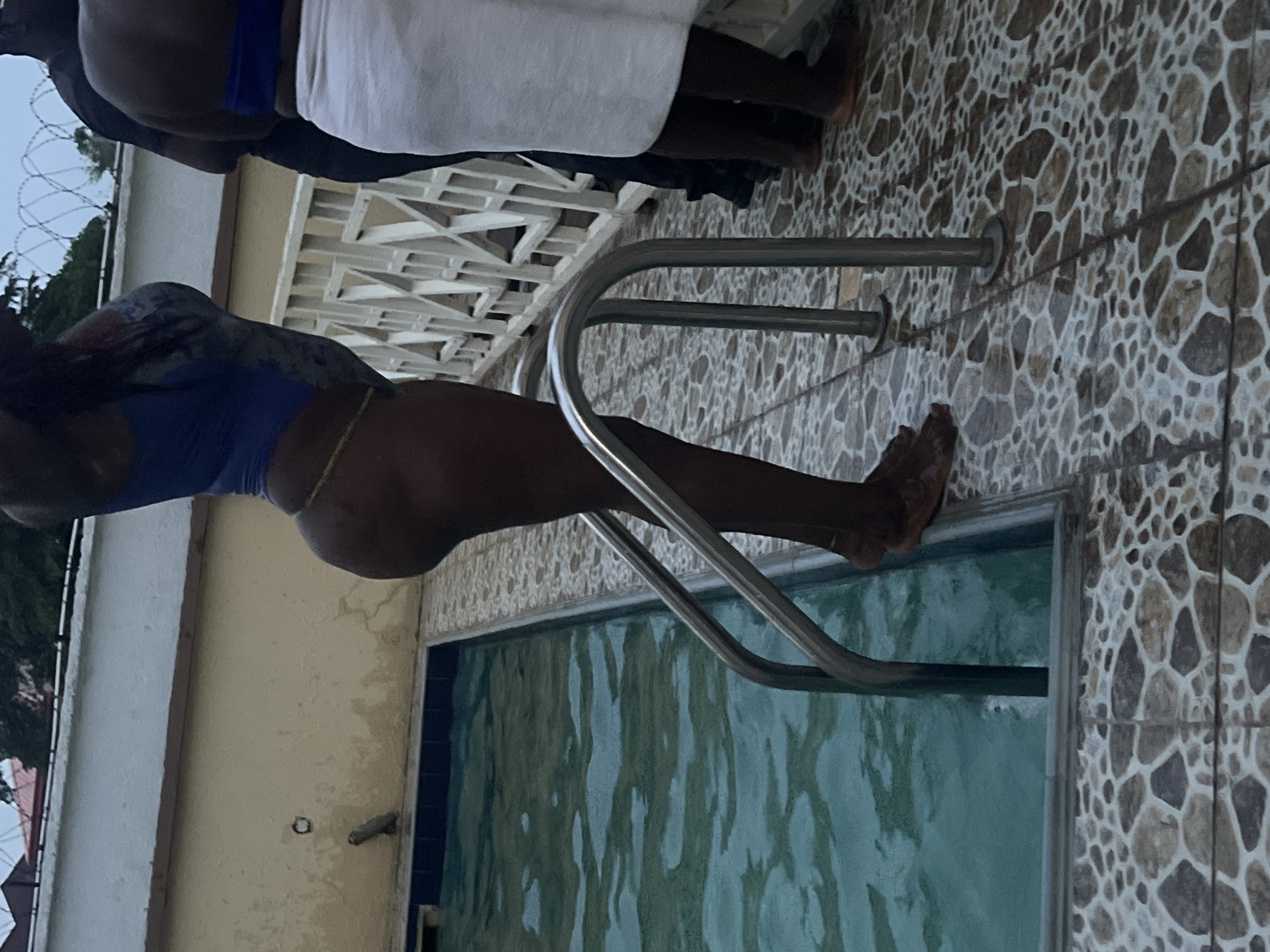 An image, rotated 90 degrees clockwise, captures an outdoor swimming pool area in Abuja, Nigeria. In the foreground, a person wearing a blue swimsuit stands with one leg in or near the water, holding onto a metal pool ladder, appearing to be entering or exiting the pool. Another individual is partially obscured by the pool ladder. Behind them, two more people are standing by the poolside; one is wrapped in a white towel, and the other wears dark clothing.
The pool itself has light blue-green water, reflecting some patterns from the surface. The area immediately surrounding the pool is paved with distinctive patterned tiles in shades of brown, grey, and white. A light-colored building wall with a decorative white fence or railing runs along one side of the pool area. In the distance, against an overcast or late afternoon sky, a barbed wire fence is visible, adding a practical detail to the setting. No discernible text is visible in the image.