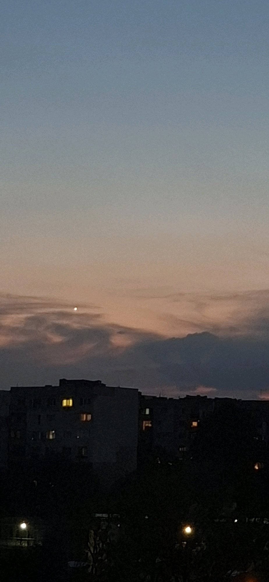 The image depicts a sky at dusk over a residential area in Sofia, Bulgaria. The sky transitions from a pale blue at the top to a warm, peachy hue lower down, interspersed with wispy clouds tinged with pink and gray. A bright, singular light source, possibly Venus or a distant planet, is visible in the upper central portion of the sky.
Below the sky, the silhouette of apartment buildings dominates the middle ground. Numerous windows are illuminated with warm, yellow light, indicating that residents are present within. The buildings are dark and featureless due to the low light conditions.
The foreground is almost entirely black, suggesting dense foliage or trees that are not illuminated. A few scattered, soft lights are visible at the very bottom of the image, possibly streetlights or lights from ground-level structures. The overall impression is of a quiet, residential evening in an urban setting.