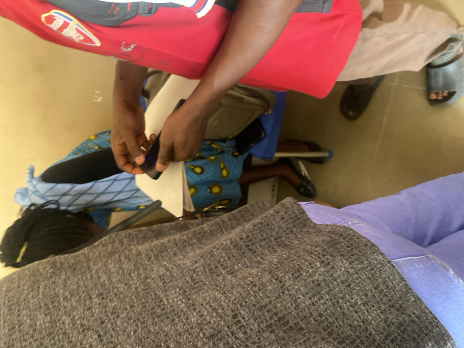 The image depicts an indoor scene in Baganakwo, Nigeria, involving three individuals. In the foreground on the left, an individual is partially visible, wearing a gray textured top and light purple trousers. To the right, another individual, wearing a red shirt with an Arsenal FC crest on the left chest and light brown trousers, is actively manipulating a dark smartphone with both hands over white sheets of paper. This individual wears dark open-toed sandals. In the background, a third individual is seated on a blue chair. This person wears a blue dress with a yellow circular pattern and a light blue patterned garment, and has dark braided hair with yellow accents. Black flat shoes are visible on their feet. A second dark smartphone rests on the blue chair beside the seated individual. The environment consists of plain, light-colored walls and a light brown floor.