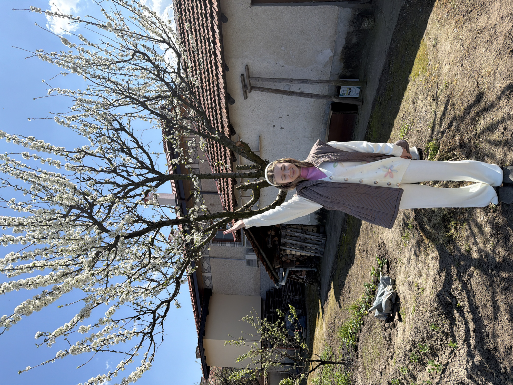 A young woman stands in a yard on a sunny day in Milevo, Bulgaria. She is wearing a white pantsuit, a purple turtleneck sweater, and a brown vest. She has a white headband in her hair. A blooming tree is to her left, and she has her right arm extended towards it. The background shows a house with a red-tiled roof. There is a pile of wood, an air conditioner, and various other structures and vegetation visible in the yard. The ground is a mix of dirt and grass.