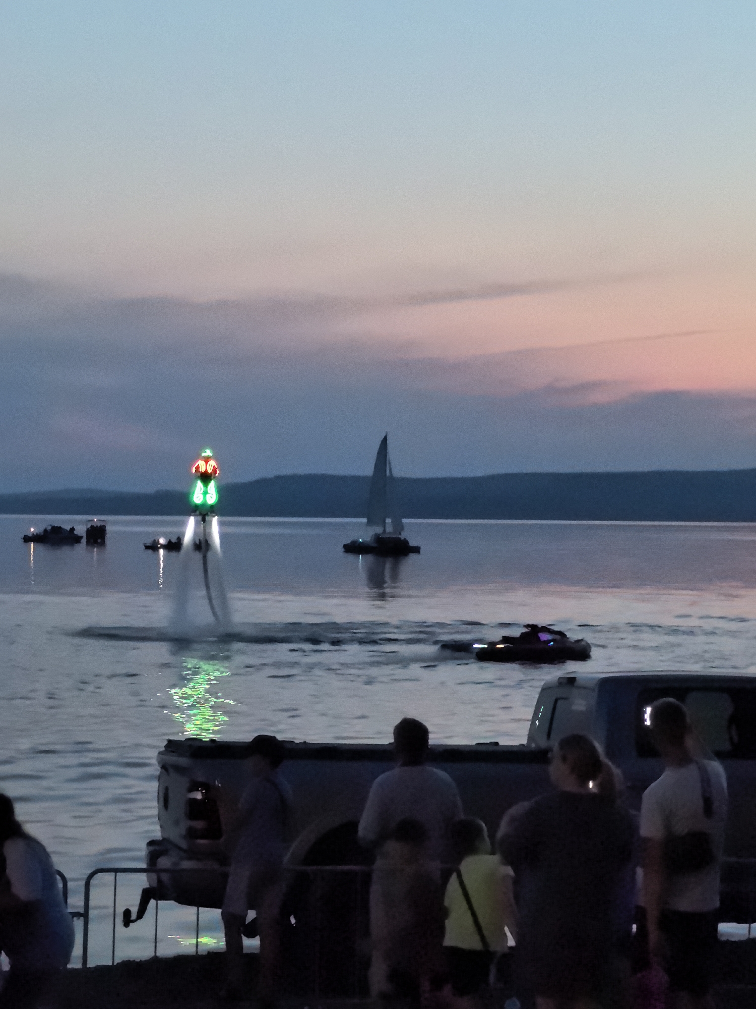 In Osa, Russia, at dusk, a group of people stand on a waterfront, watching a water show. The sky is painted with hues of blue, pink, and orange, transitioning into twilight. The calm lake reflects the soft colors of the sky. In the distance, a sailboat glides across the water. A jet ski is visible in the foreground, its lights illuminating the water. The main subject of the scene is a person on a flyboard, illuminated by neon green and red lights, soaring above the water, creating a dramatic spray. Nearby, several boats are anchored, and a pickup truck is parked on the shore, with a few spectators near it. The overall atmosphere is one of evening entertainment and community gathering by the water.