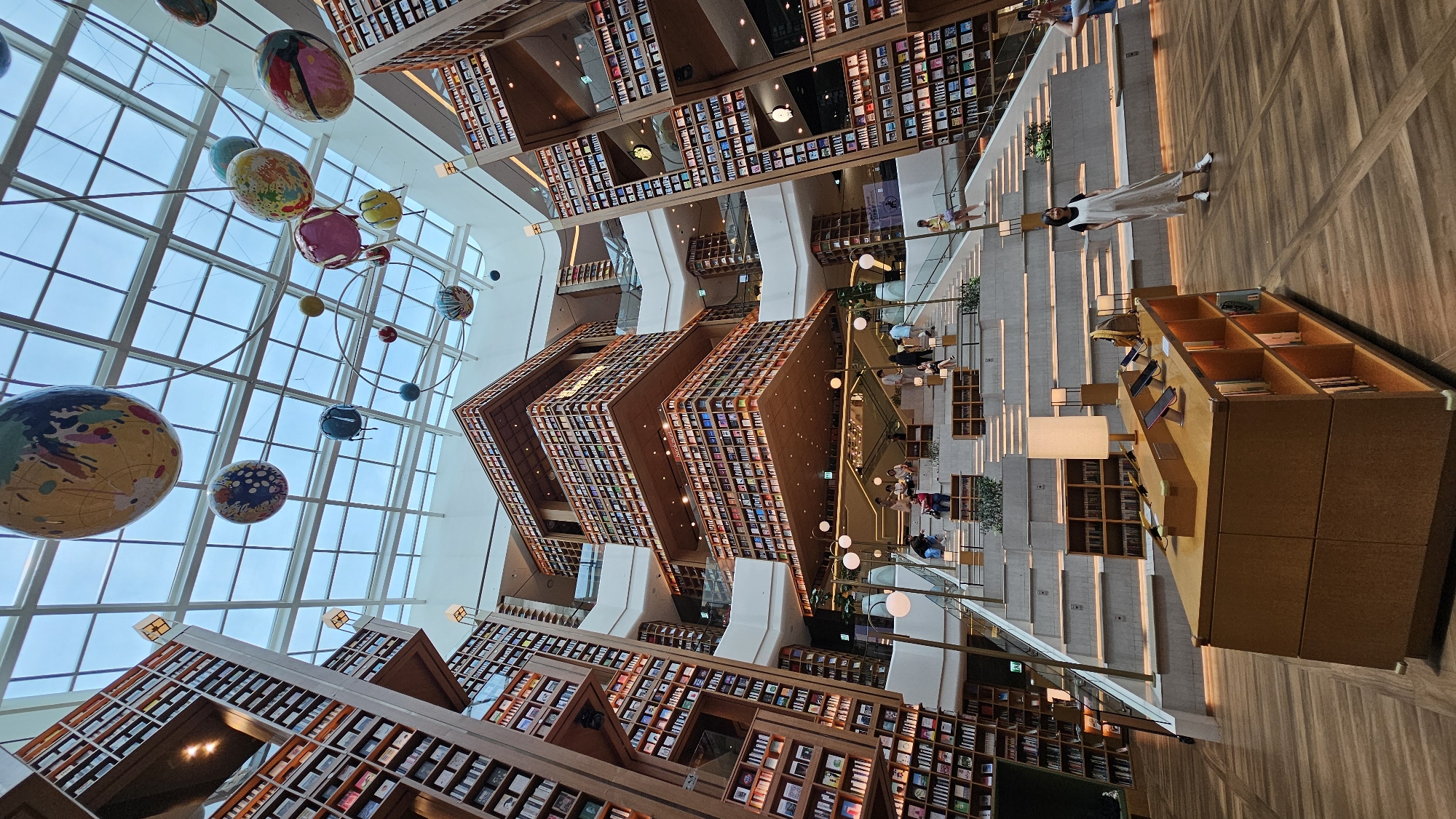 This is an interior photograph of a multi-story library with a large, atrium-like space. The library features towering bookshelves that extend upwards along the walls, filled with numerous books. Suspended from the transparent ceiling, which appears to be a skylight or glass roof, are several large, spherical decorative objects resembling planets, some adorned with colorful patterns. A grand staircase made of tiered concrete or stone steps with integrated lighting ascends through the center of the space. Small groups of people are visible on the stairs and at various levels of the library, some appearing to browse or walk. In the foreground, a wooden display table is set up, holding several electronic devices and some books. A woman in a white dress stands to the right of the display table, and another person on the far right appears to be taking a photo. The overall atmosphere suggests a modern and architecturally impressive public space. This library is likely Starfield Library located in COEX Mall, Gangnam-gu, Seoul, South Korea, not Suwon-si.