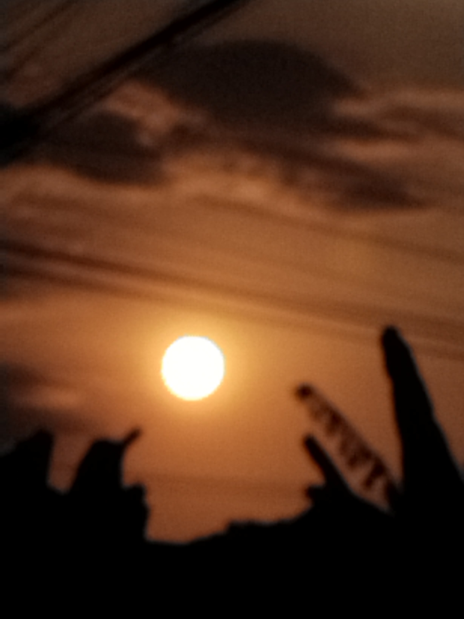 A silhouette of a sunset is visible in the image. The sun is a bright, glowing orb, positioned in the center left of the frame. The sky is a warm orange, with darker clouds scattered throughout. In the foreground, there are dark silhouettes of what appear to be people or objects, with two of them making a peace sign with their fingers. Overhead, there are power lines. The image is slightly blurred, giving it a dreamy quality.